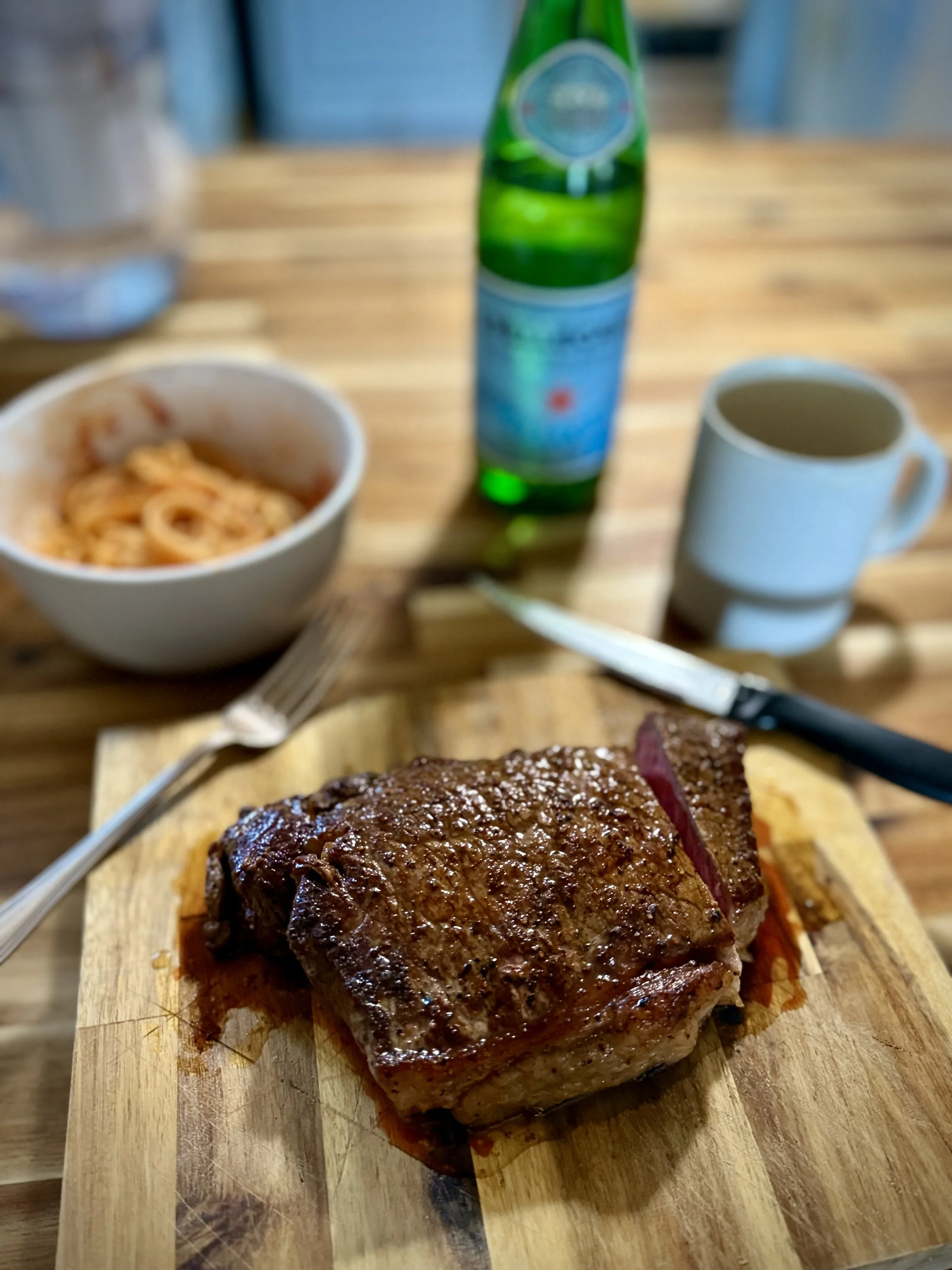 Cooked steak on a wooden board with a fork and knife, a bowl of pasta, a green bottle, and a white mug on a wooden table.