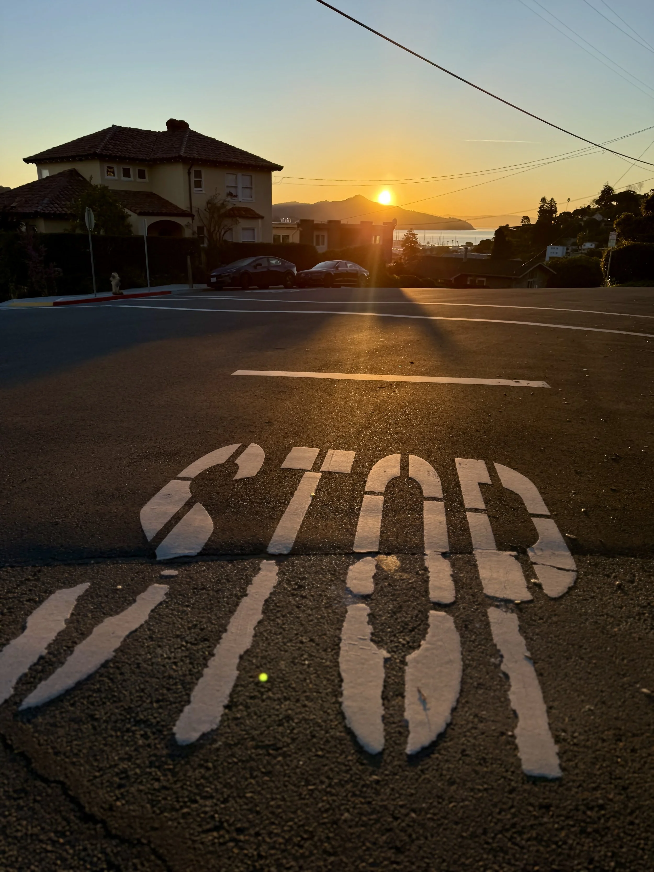Street view with "STOP" painted on the pavement, residential houses, parked cars, sunset over distant hills, and power lines overhead.