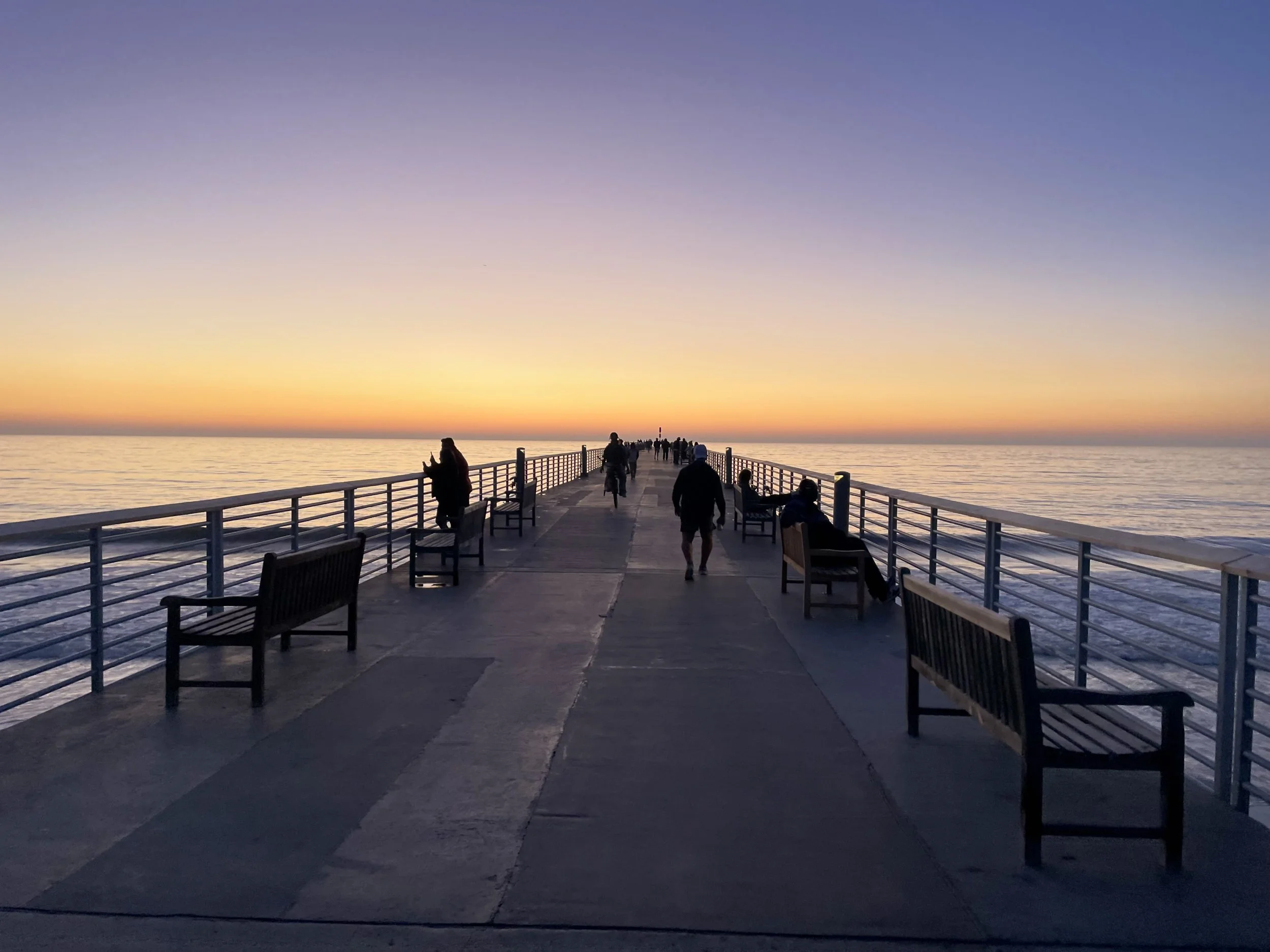 People walking on a pier at sunset with benches and railing, overlooking the ocean and a colorful sky.