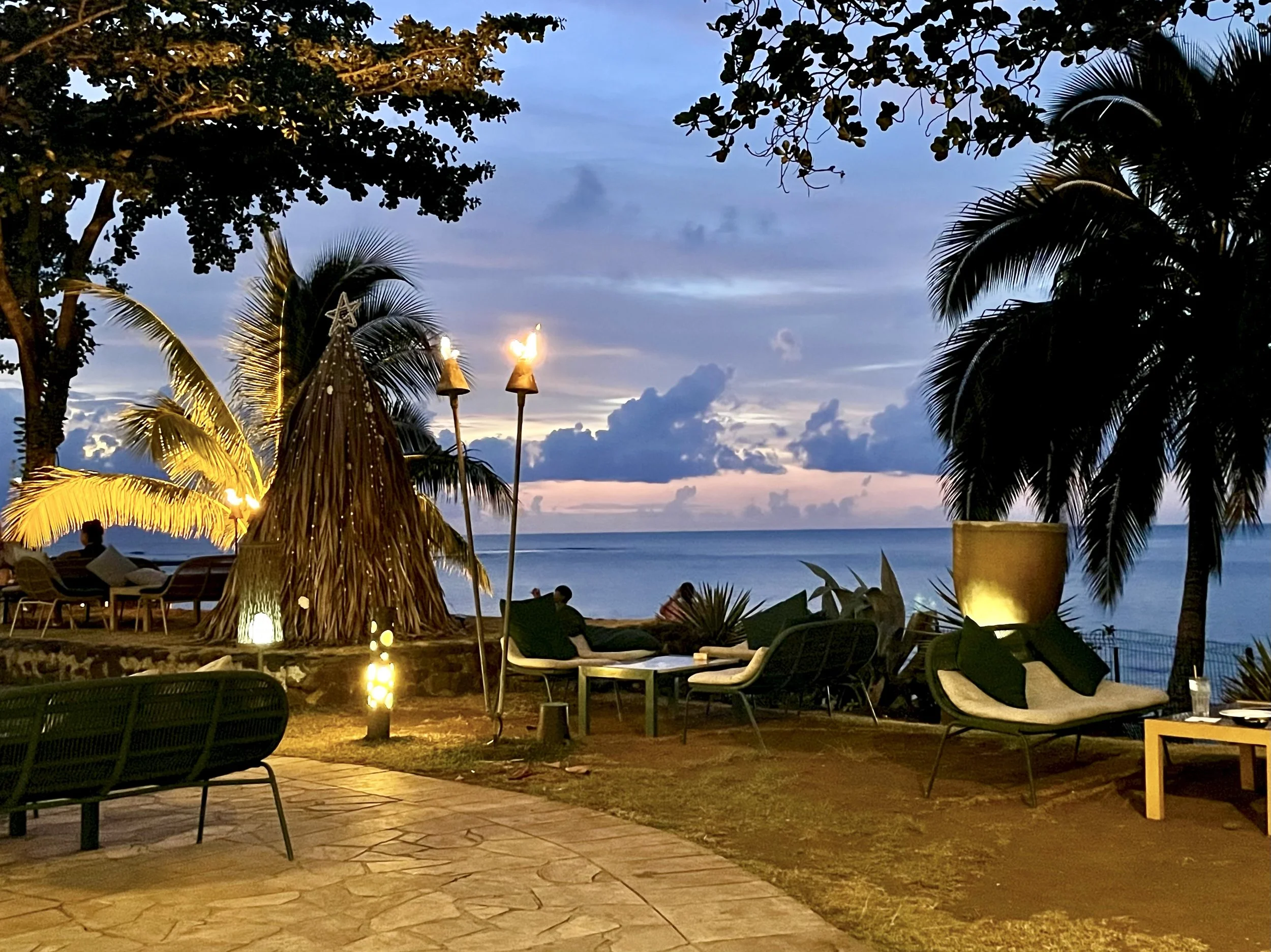 Outdoor tropical seating area overlooking the ocean at sunset, surrounded by palm trees and lit tiki torches.