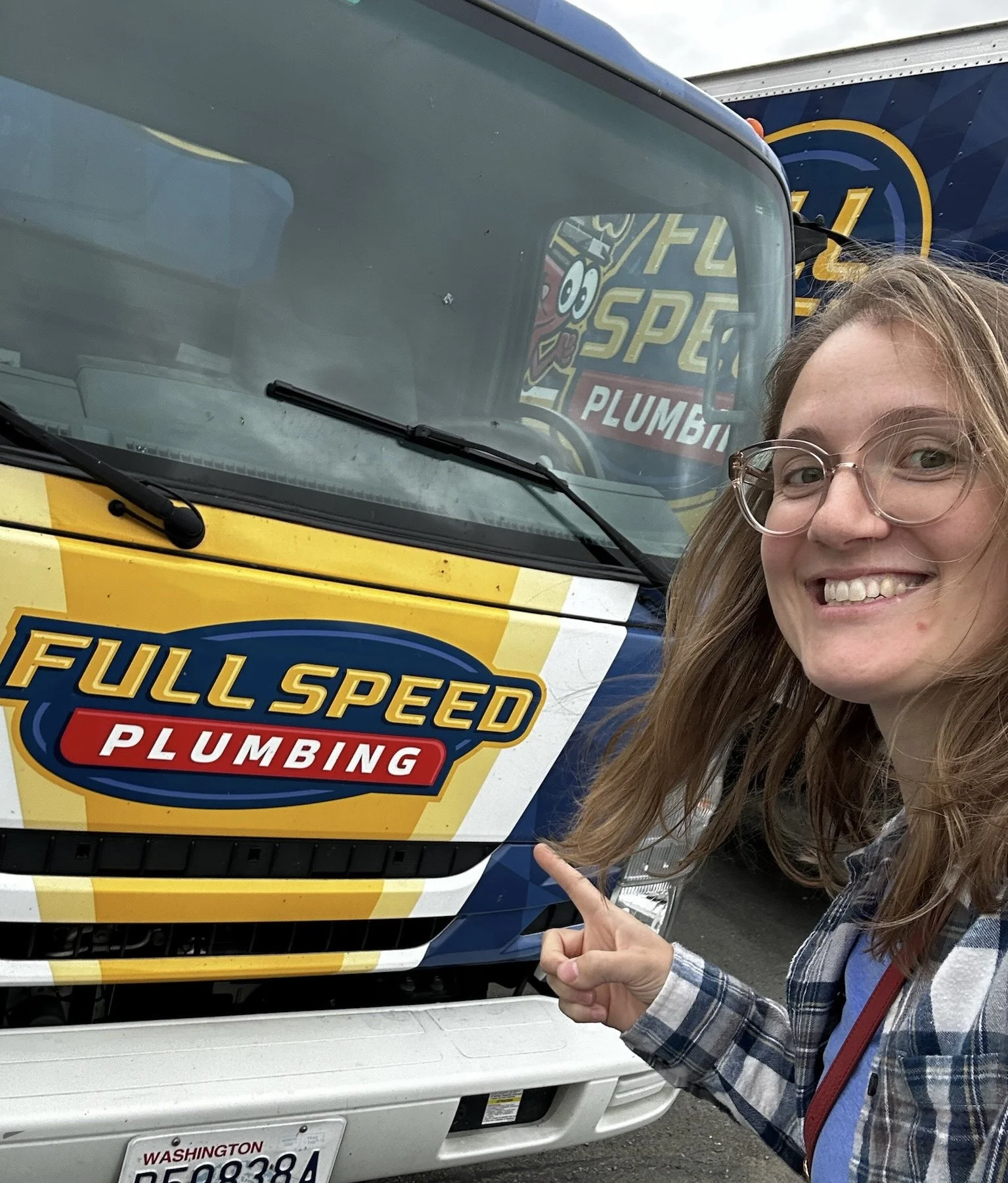 A smiling woman with glasses pointing at a truck with a 'Full Speed Plumbing' logo on its front. The truck has a yellow and blue color scheme, and there is a person reflected in the windshield.