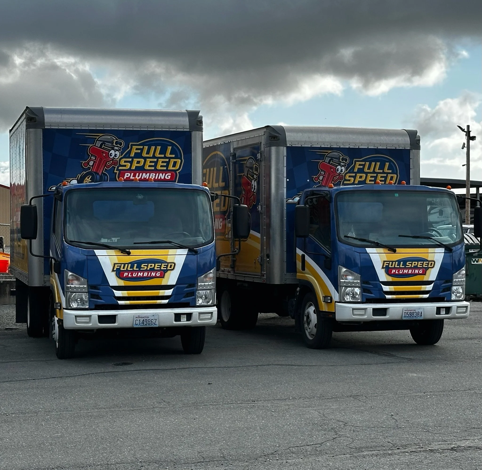 Two full-speed plumbing delivery trucks parked side by side outdoors under cloudy sky.