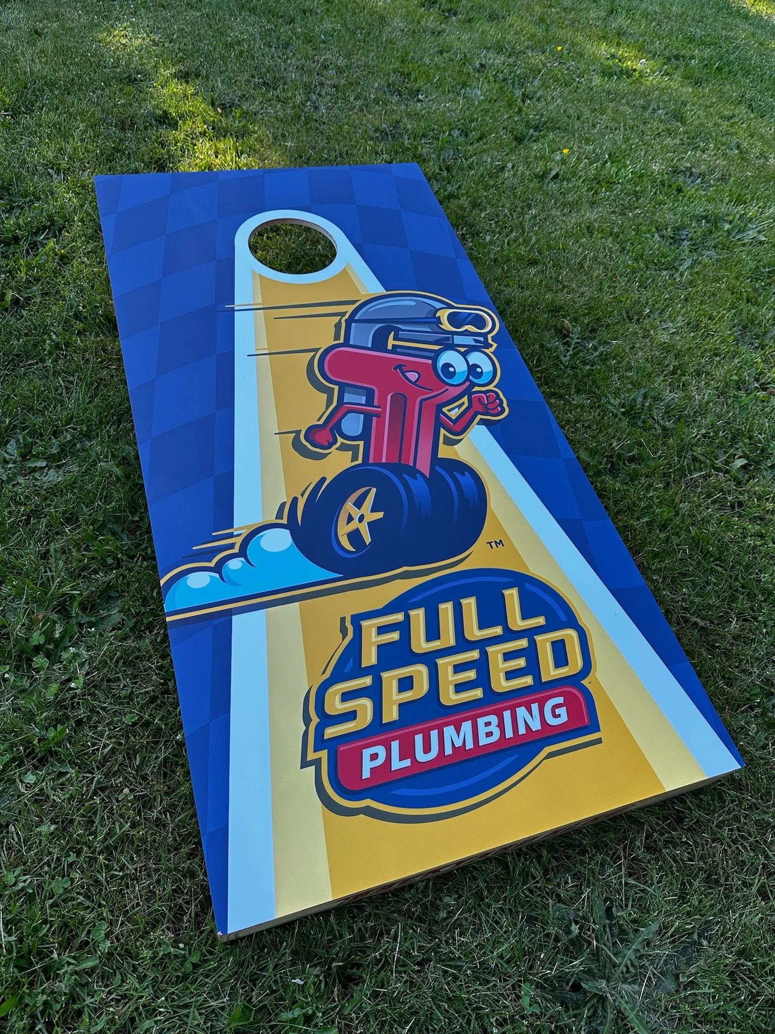 Cornhole game board with a cartoon character as a rocket and the text 'Full Speed Plumbing' on it, placed on grass.