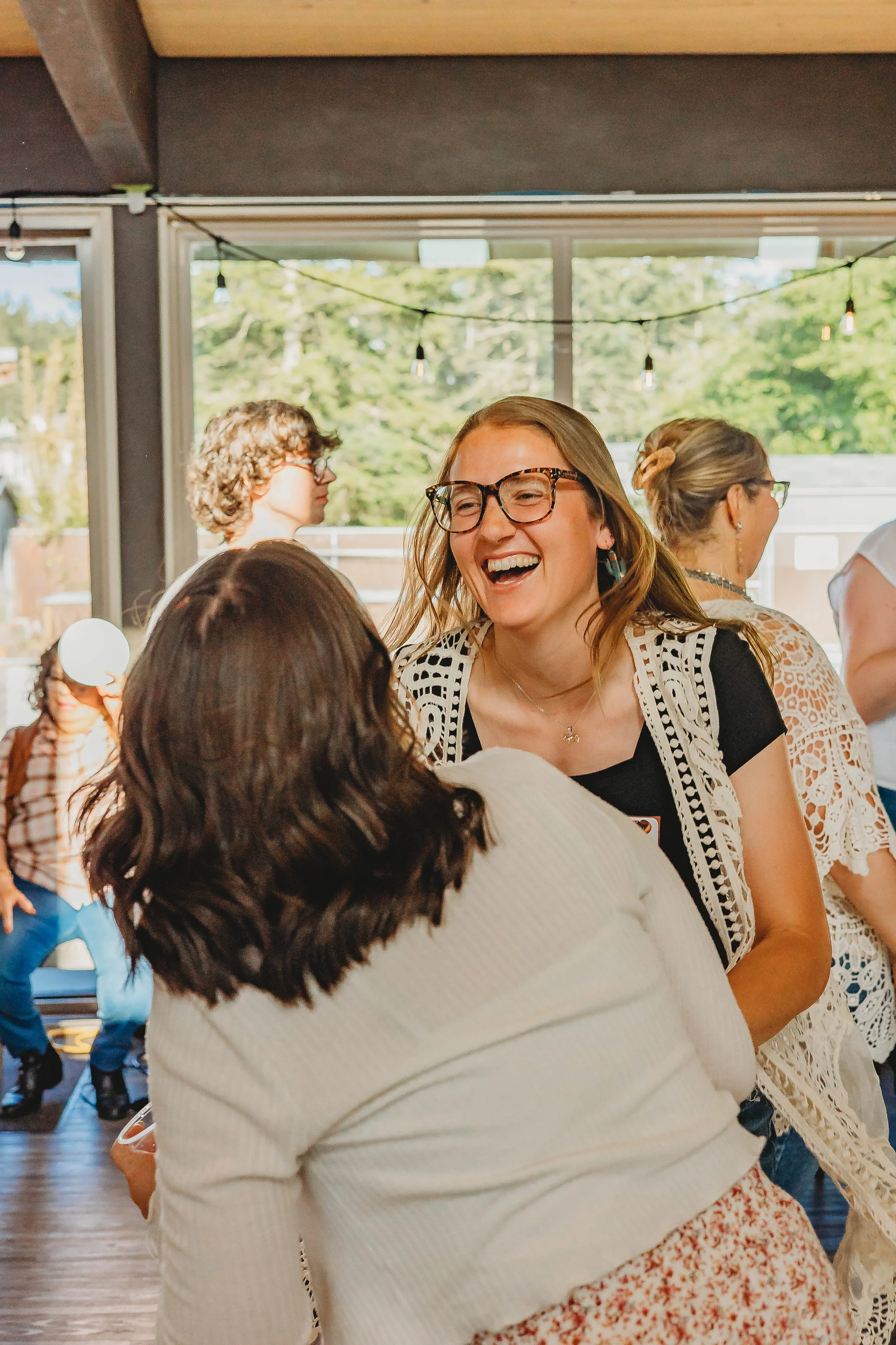 People at an indoor gathering, smiling, laughing, and talking, with string lights hanging from the ceiling and large windows showing trees outside.