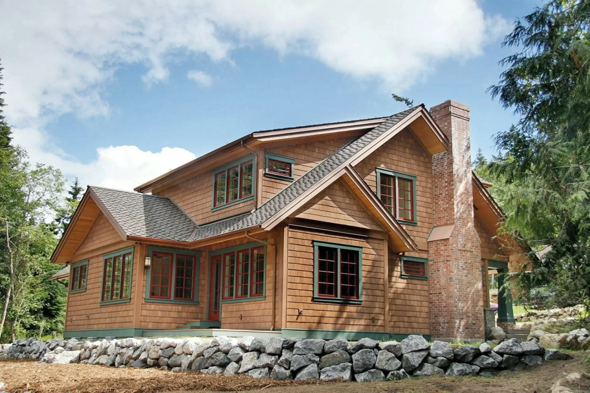 A multi-story house with wooden siding, multiple windows with green frames, and a brick chimney, surrounded by trees and a stone wall in a lush, green landscape under a partly cloudy sky.