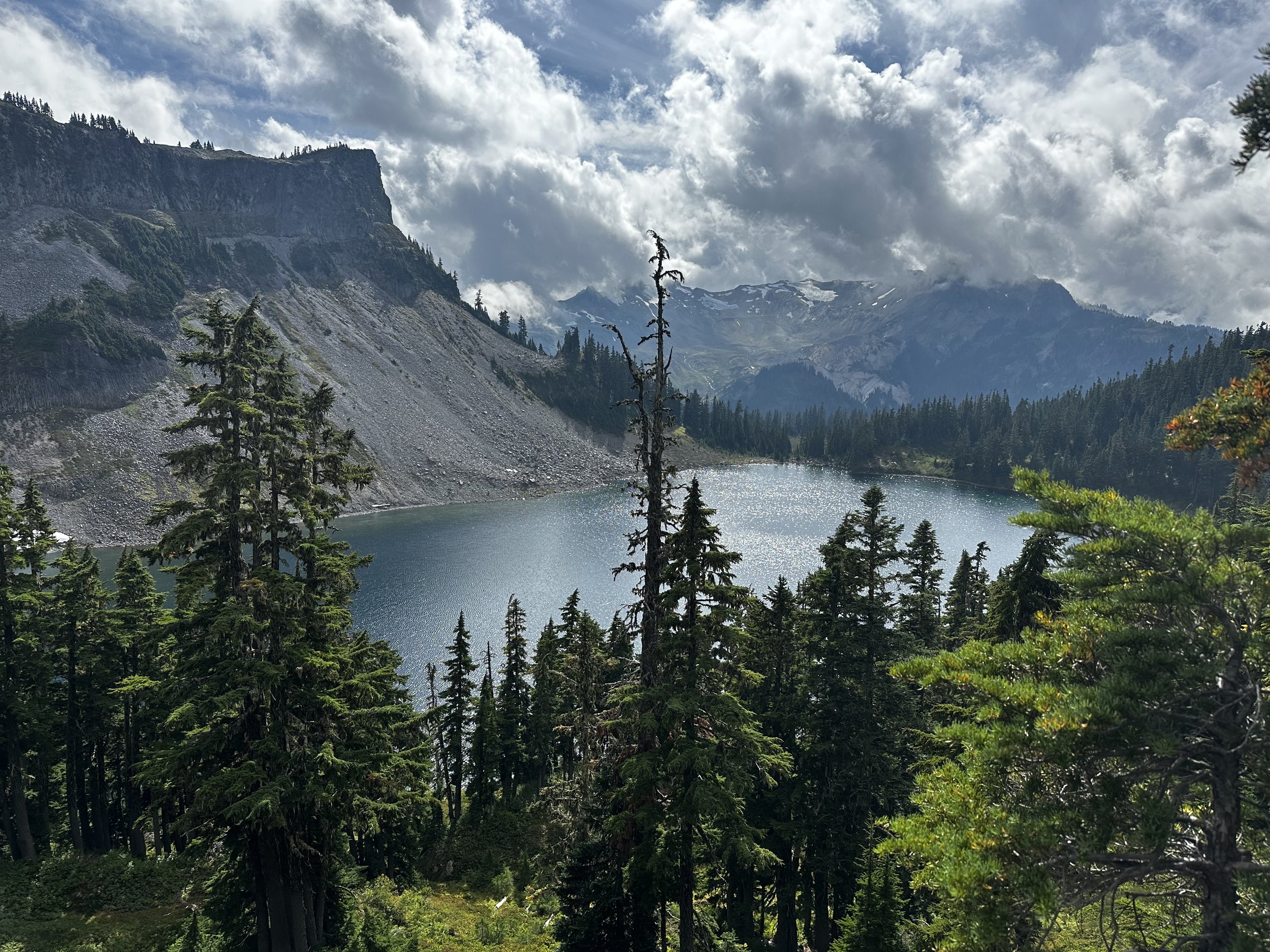 Scenic view of a mountain lake surrounded by dense evergreen trees, with rocky mountain slopes and snow-capped peaks in the background under a partly cloudy sky.