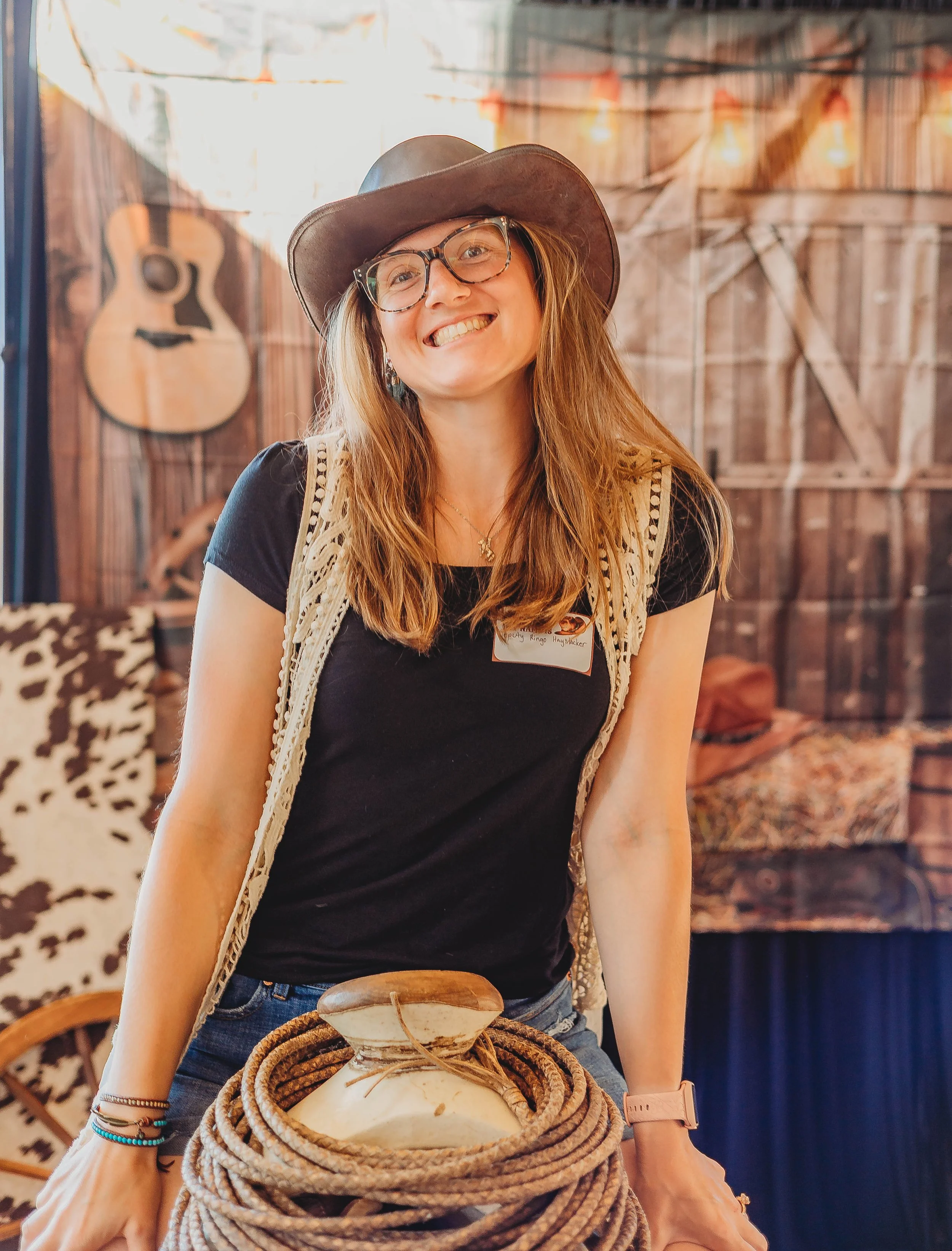 A smiling woman with glasses, wearing a cowboy hat, a black T-shirt, and a cream-colored vest, sitting on a saddle in a rustic setting with wooden walls and musical instruments in the background.