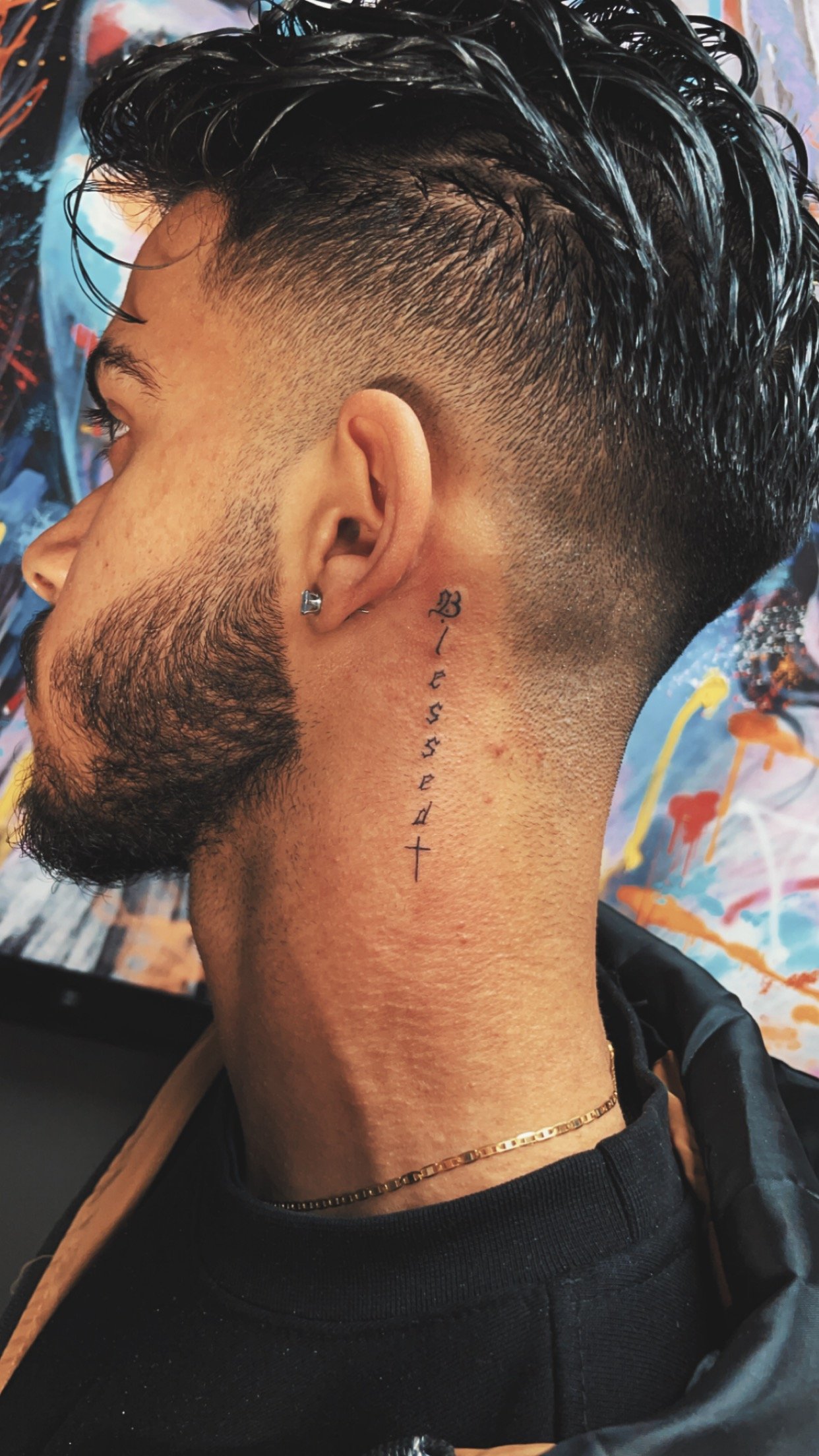Side view of a person with a 'Blessed' tattoo in cursive on the neck, adorned with an earring and a chain necklace, against a colorful abstract background.