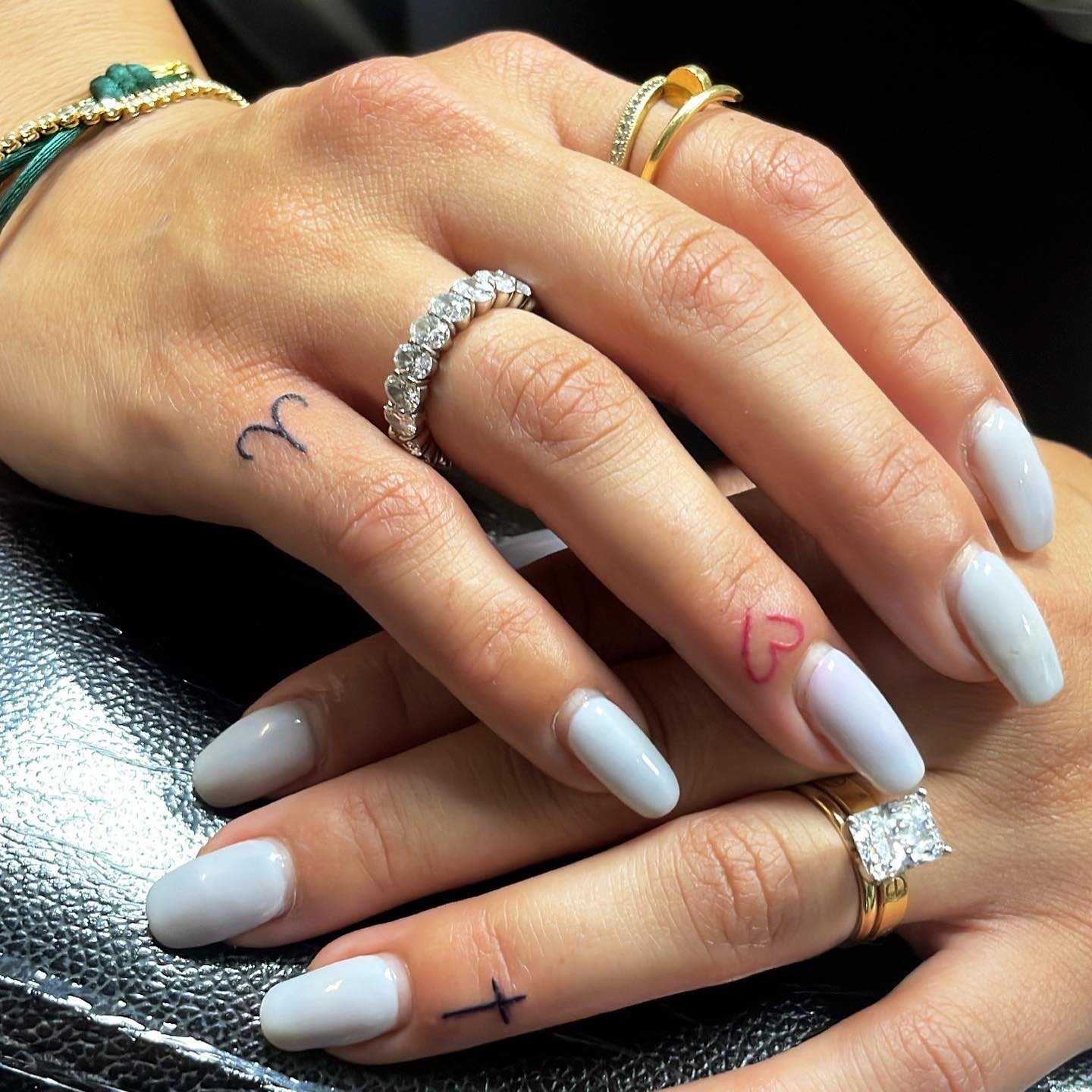 Close-up of two hands with light gray manicured nails, adorned with gold and diamond rings. Small tattoos on fingers include a heart, a symbol resembling Aries, and a cross. Hands rest on a textured black surface.