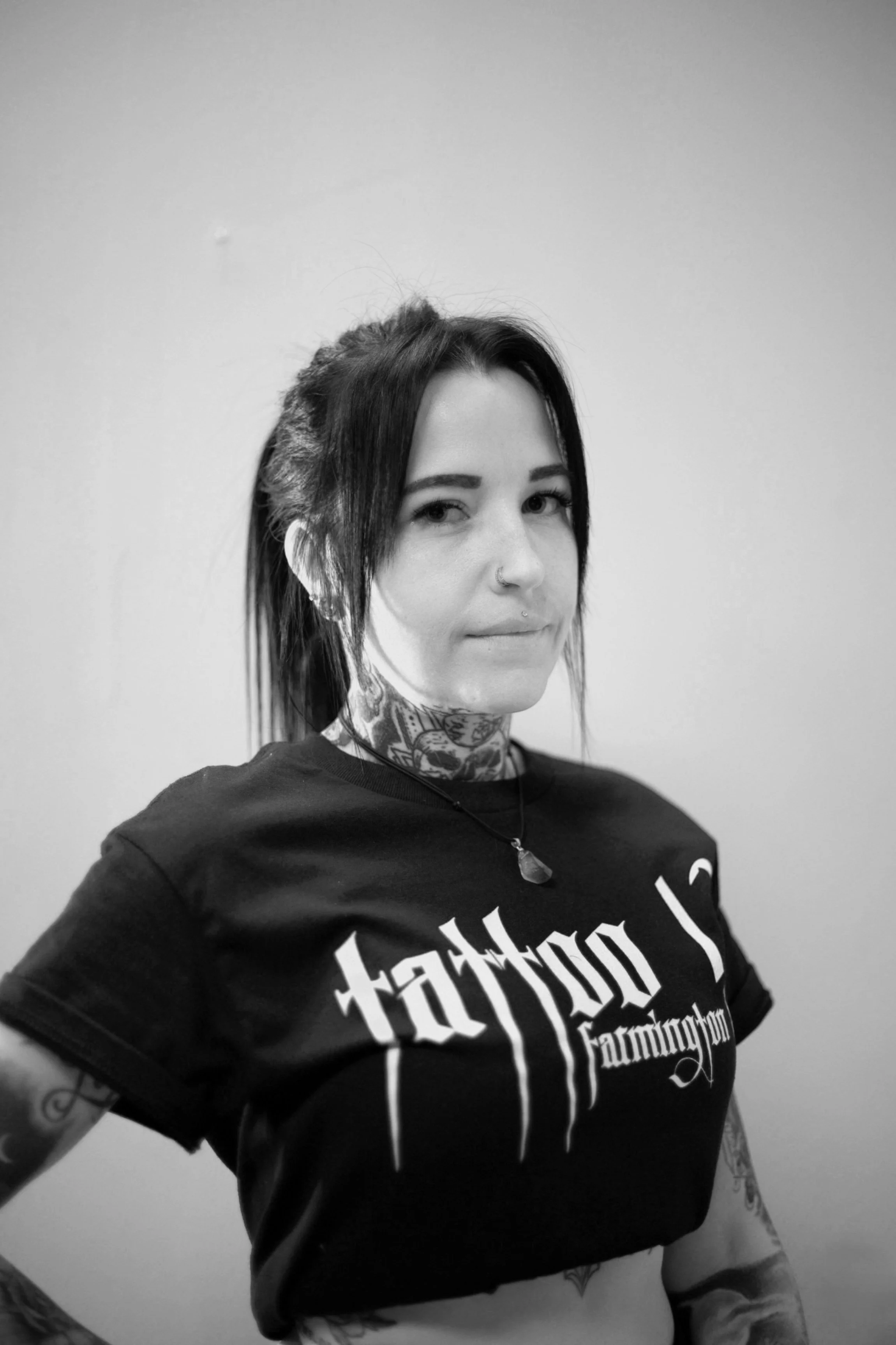 Black and white photo of a person with tattoos wearing a "Tattoo 13 Farmington" T-shirt and a necklace, with a nose ring.