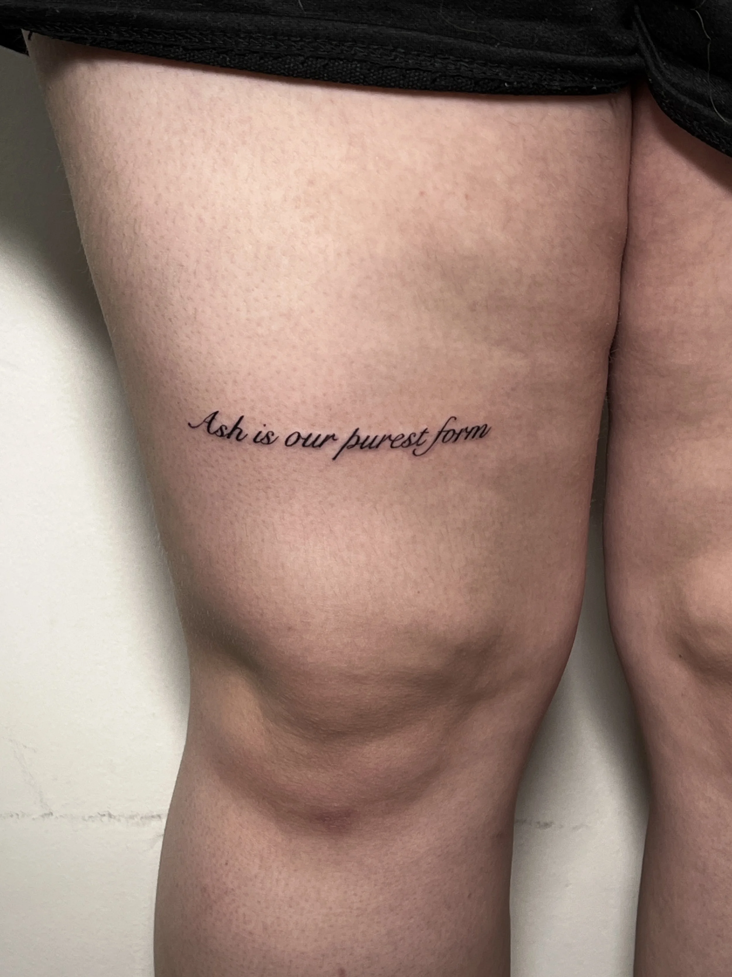 Close-up of a person's upper thigh and hip with a black tattooed quote, wearing black clothing.