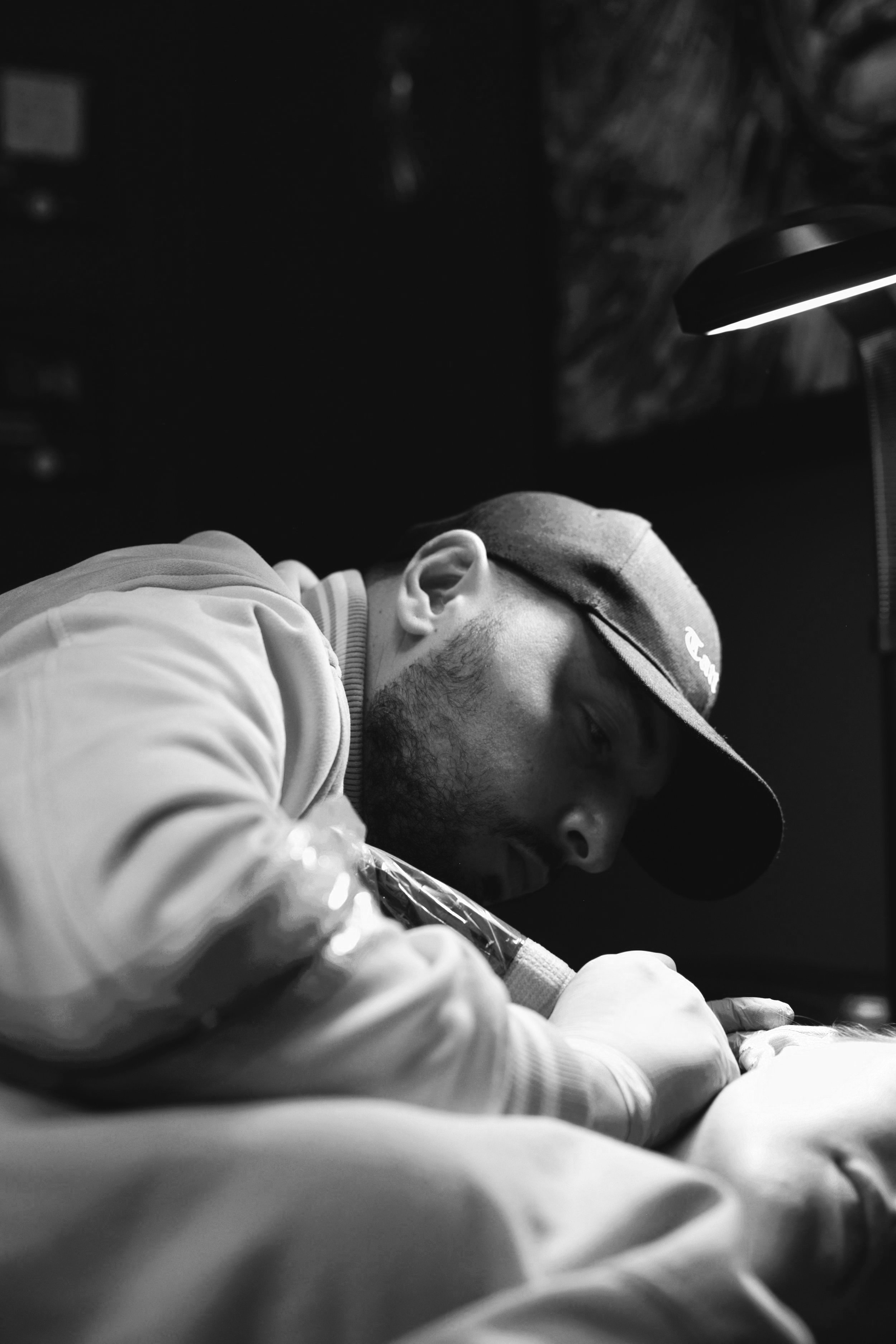 Black and white photo of a tattoo artist working intently on a client under a lamp.