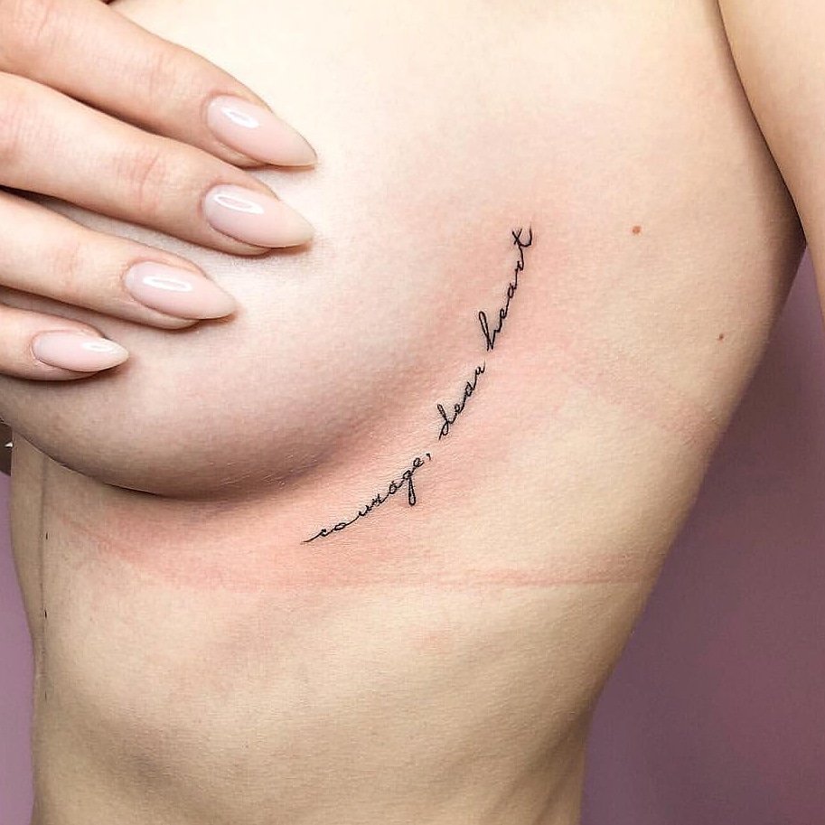 Close-up of a script tattoo reading "courage, dear heart" under a breast, with a hand covering part of the breast and nails painted in a natural color.