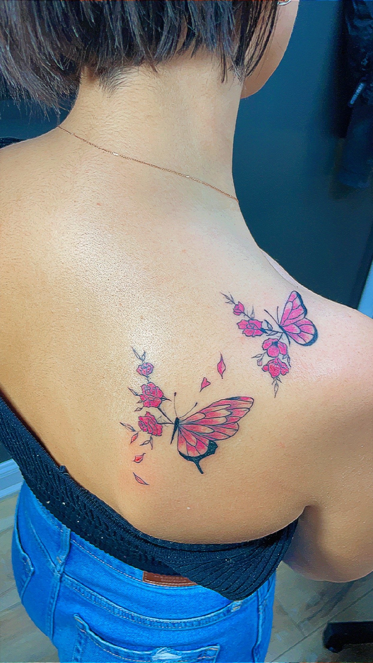 Woman with short hair showing a tattoo on her back, featuring pink butterflies and flowers.