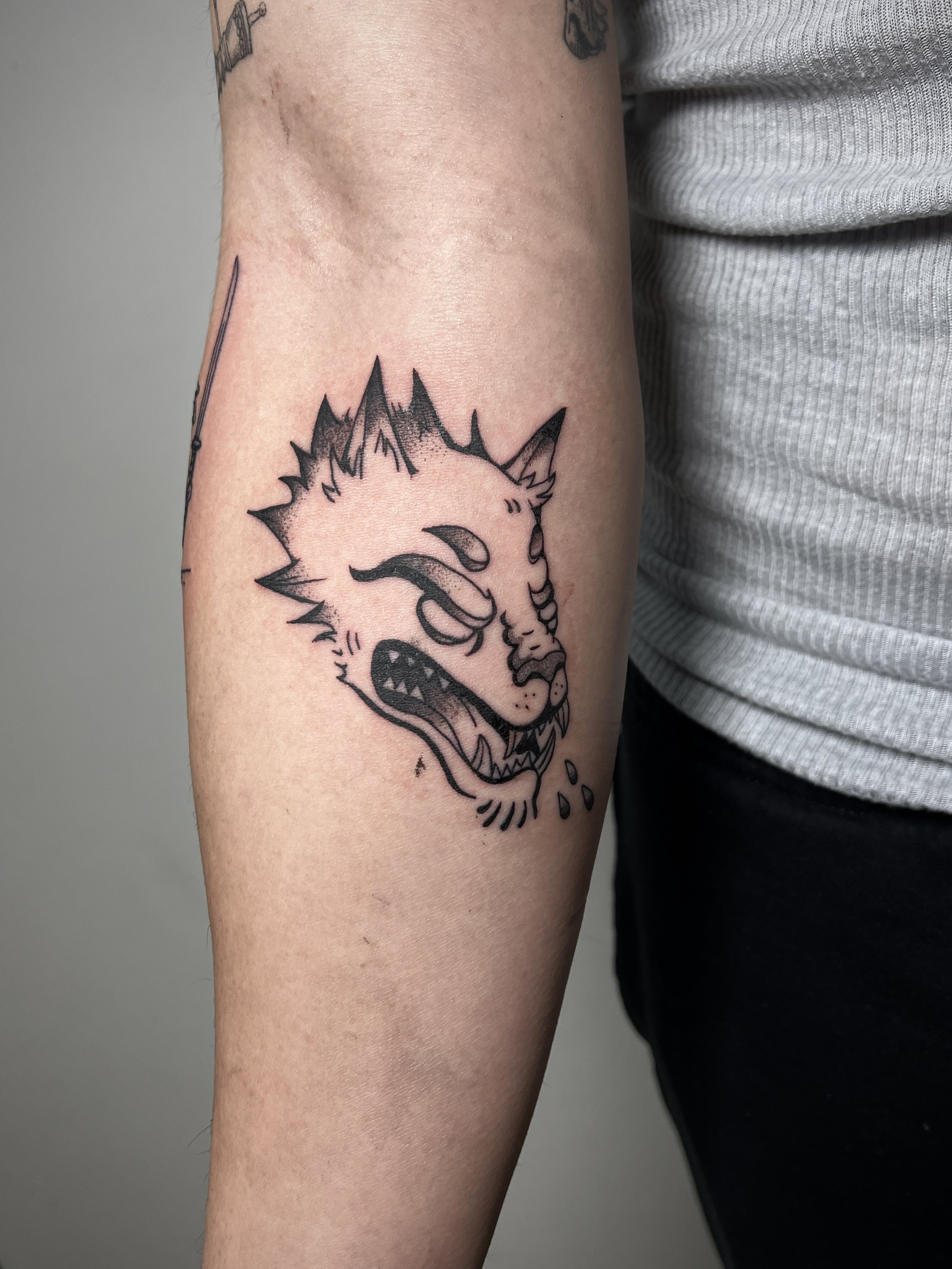 A person's upper arm with a black tattoo of a snarling wolf's face with spiky fur, and some other tattoos visible in the background.