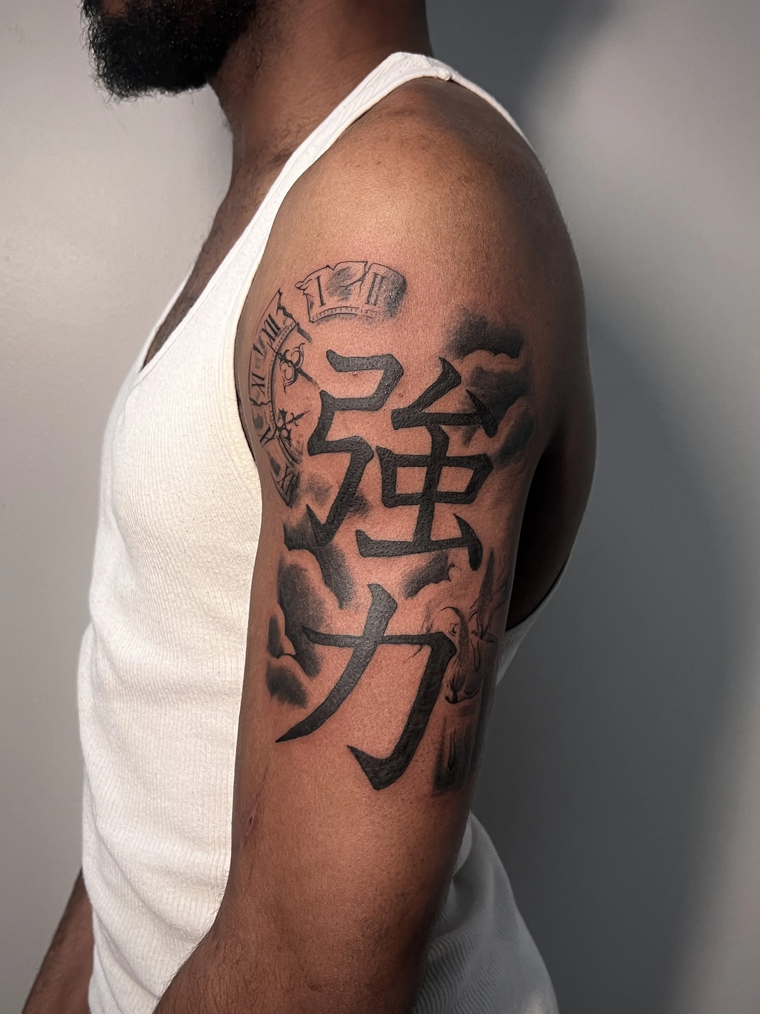 A person's upper arm with a tattoo of a clock and large Japanese characters, wearing a white tank top.