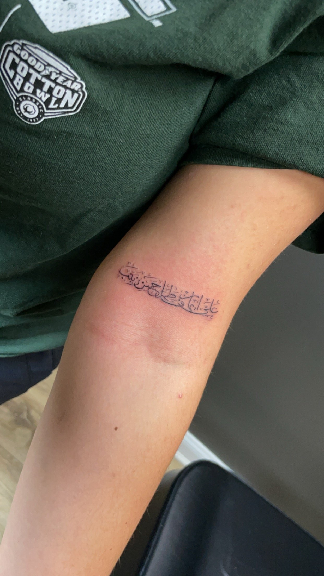 Person's arm with a tattoo in Arabic script, wearing a green shirt with "Goodyear Cotton Bowl" logo.