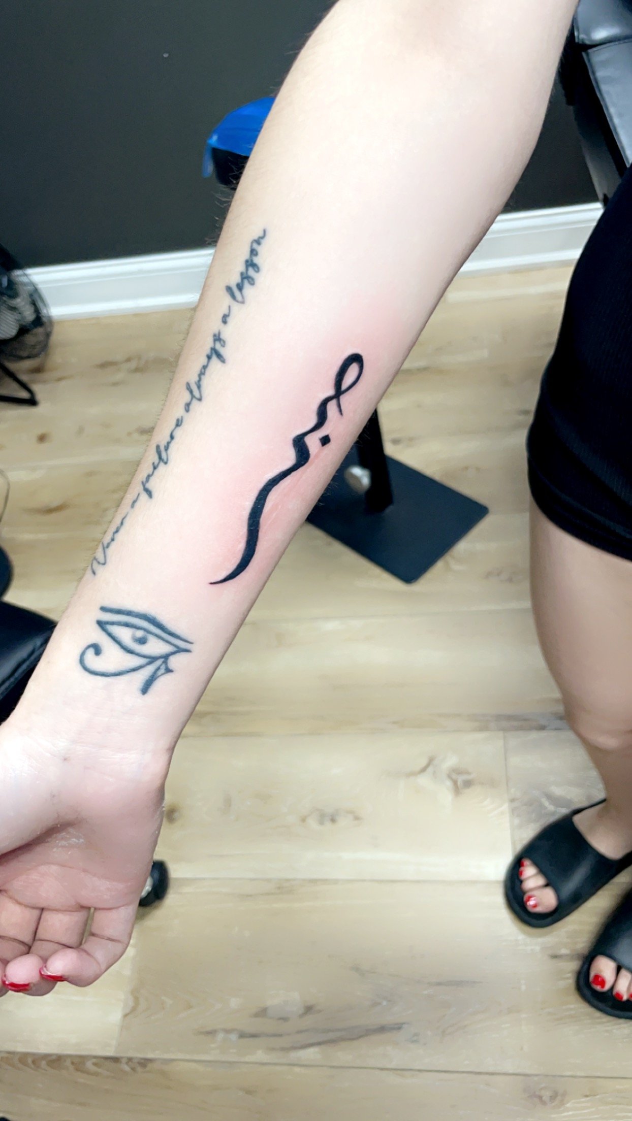 Forearm tattoos with an Eye of Horus and Arabic script on a person standing on a wooden floor.