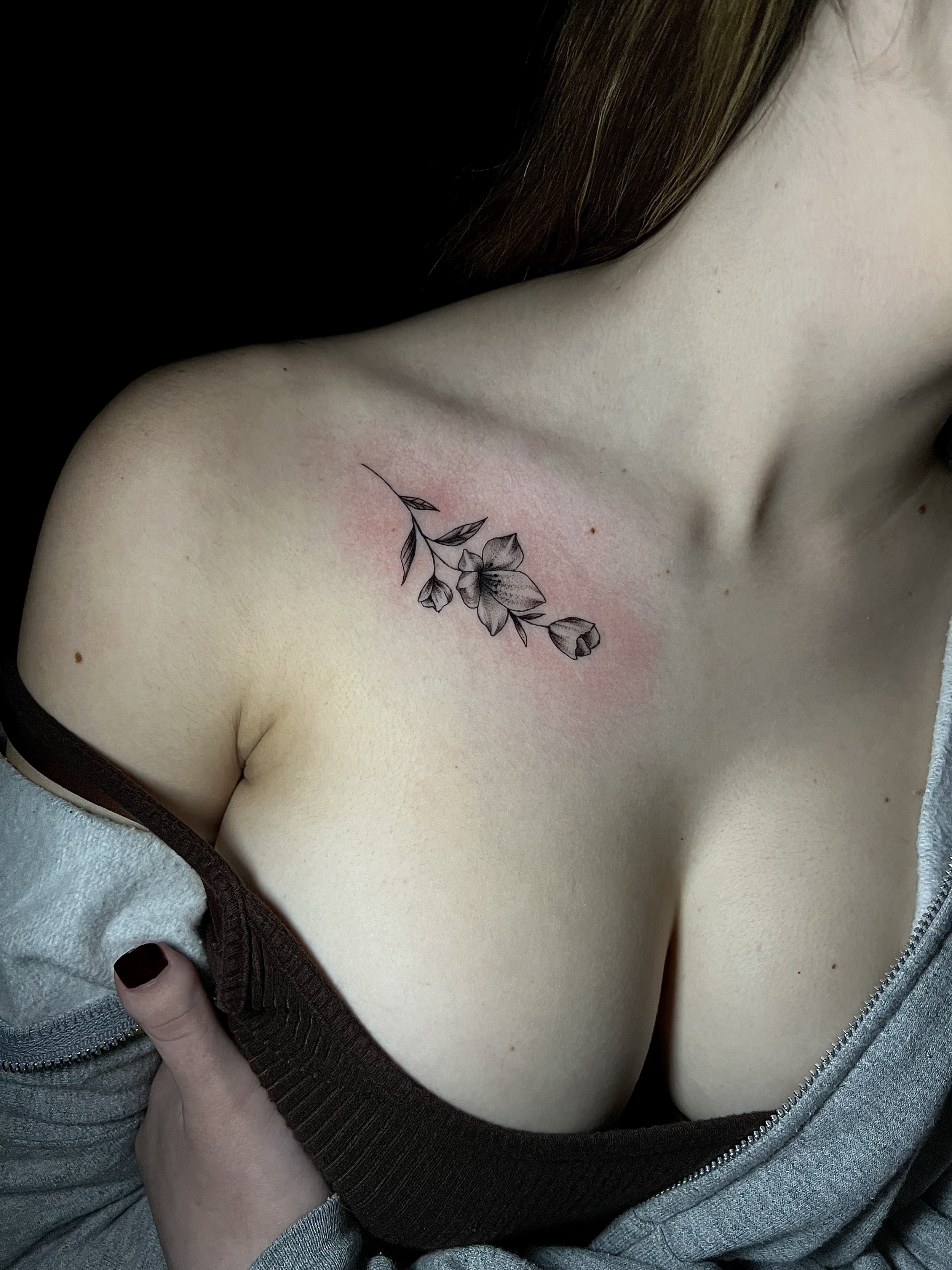 Close-up of a floral tattoo on a woman's collarbone, with a brown strap partially off her shoulder and wearing a gray zip-up jacket.