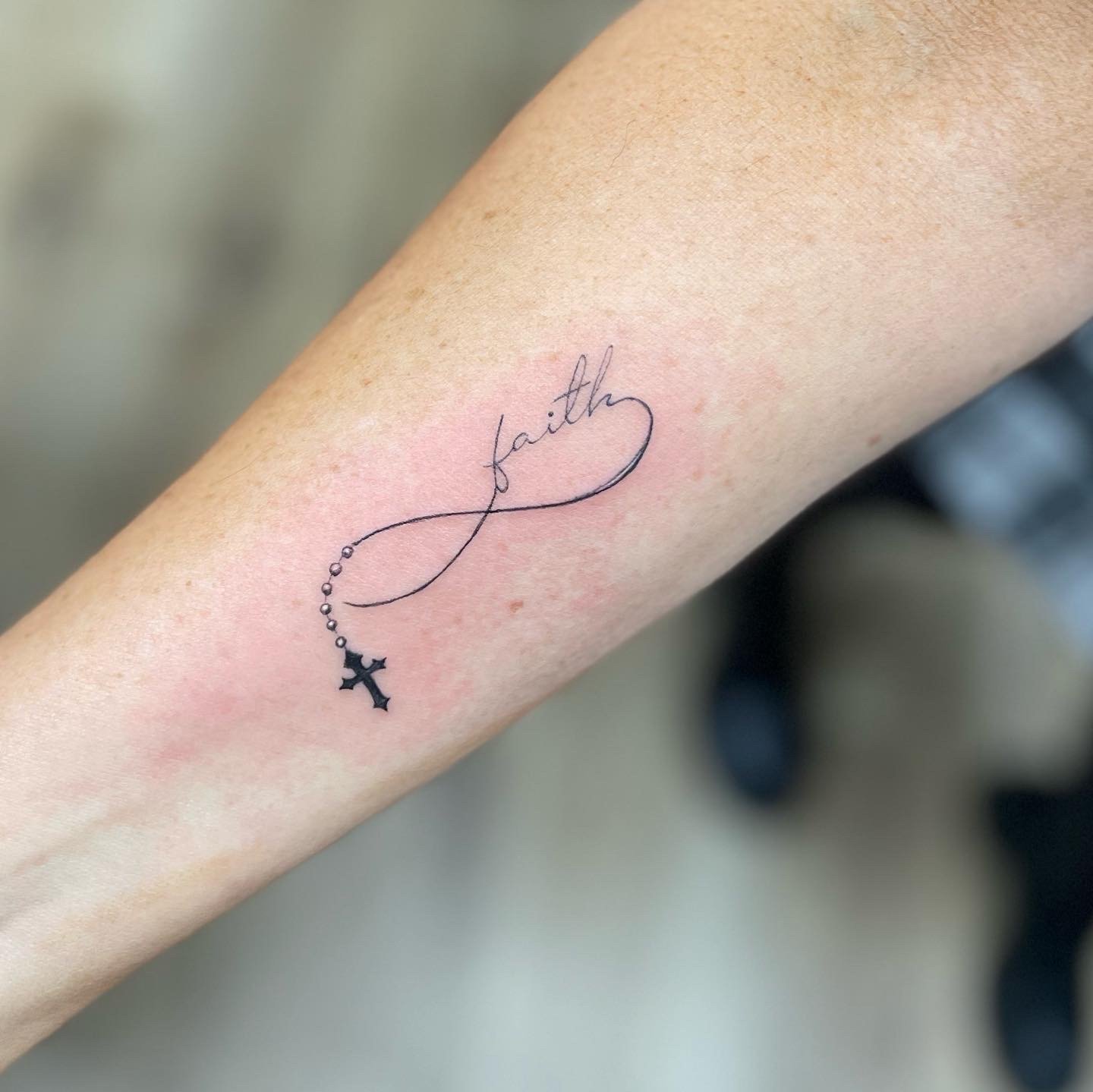 Tattoo of the word 'faith' with rosary beads and cross on an arm.