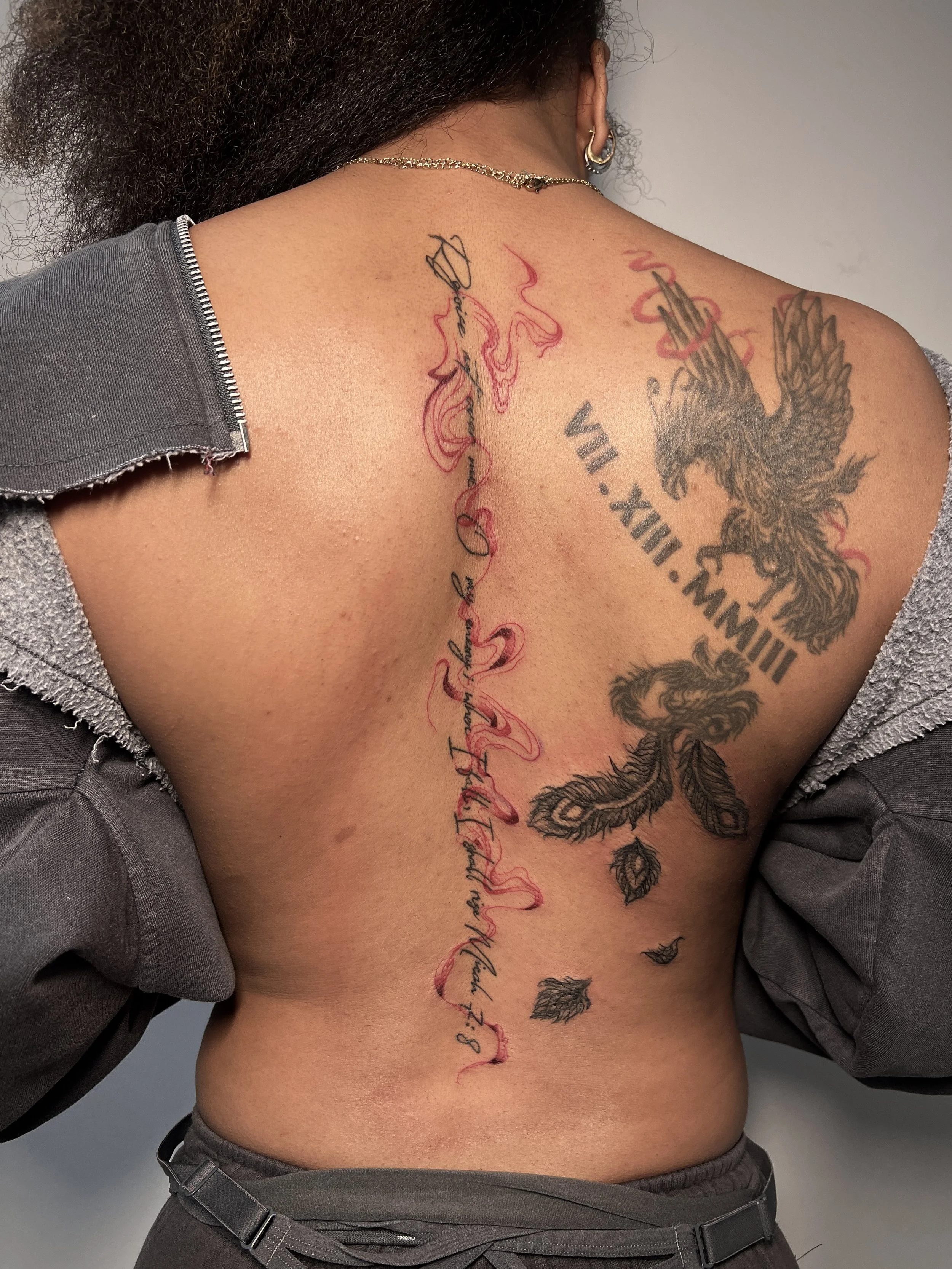 A person's back decorated with multiple tattoos. Down her spine is text with abstract red smoke swirling through the script.