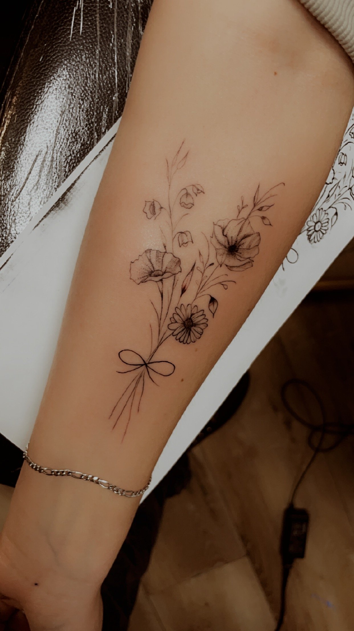 Floral tattoo on forearm with line art design of flowers and leaves tied with a ribbon.