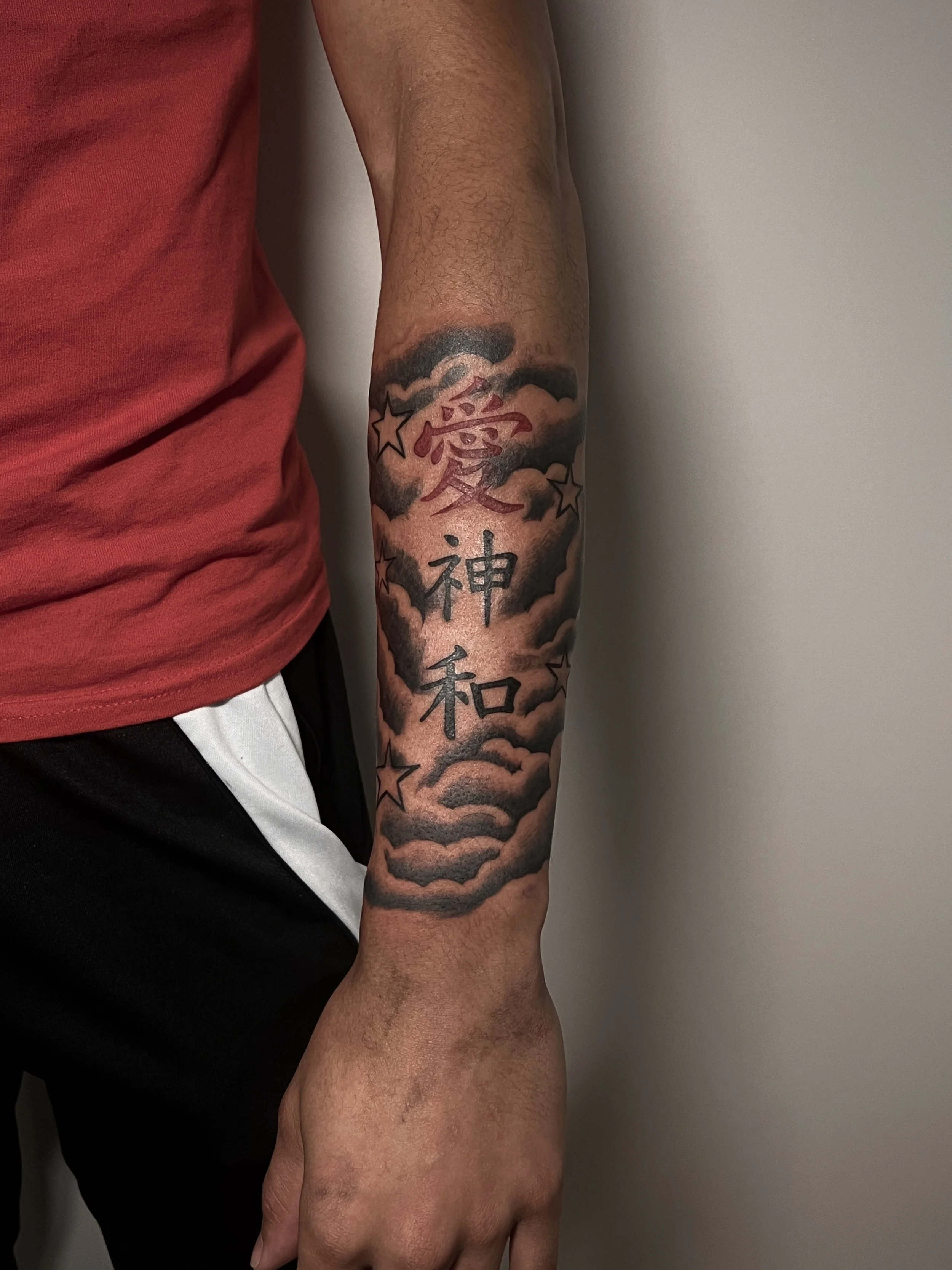 A person's arm with a tattoo featuring clouds, stars, and Japanese characters, wearing a red shirt and black shorts, standing against a plain wall.