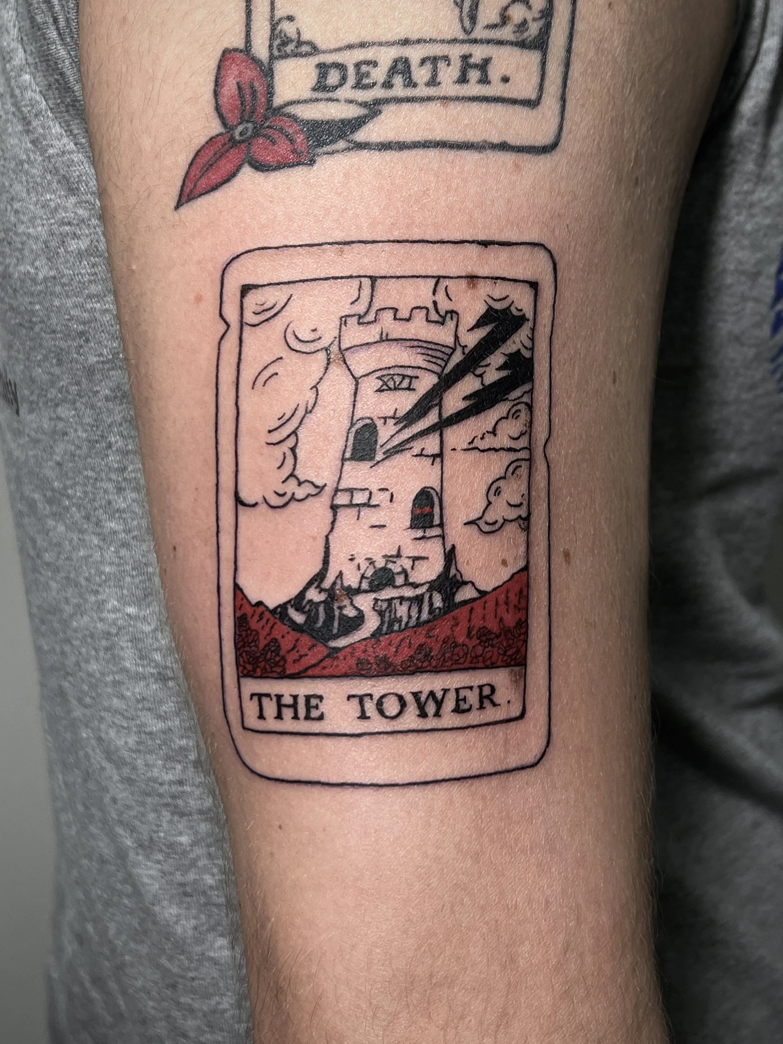 A tattoo designed to resemble the Tarot card 'The Tower,' featuring a tower with lightning and clouds, and the words 'The Tower' at the bottom.