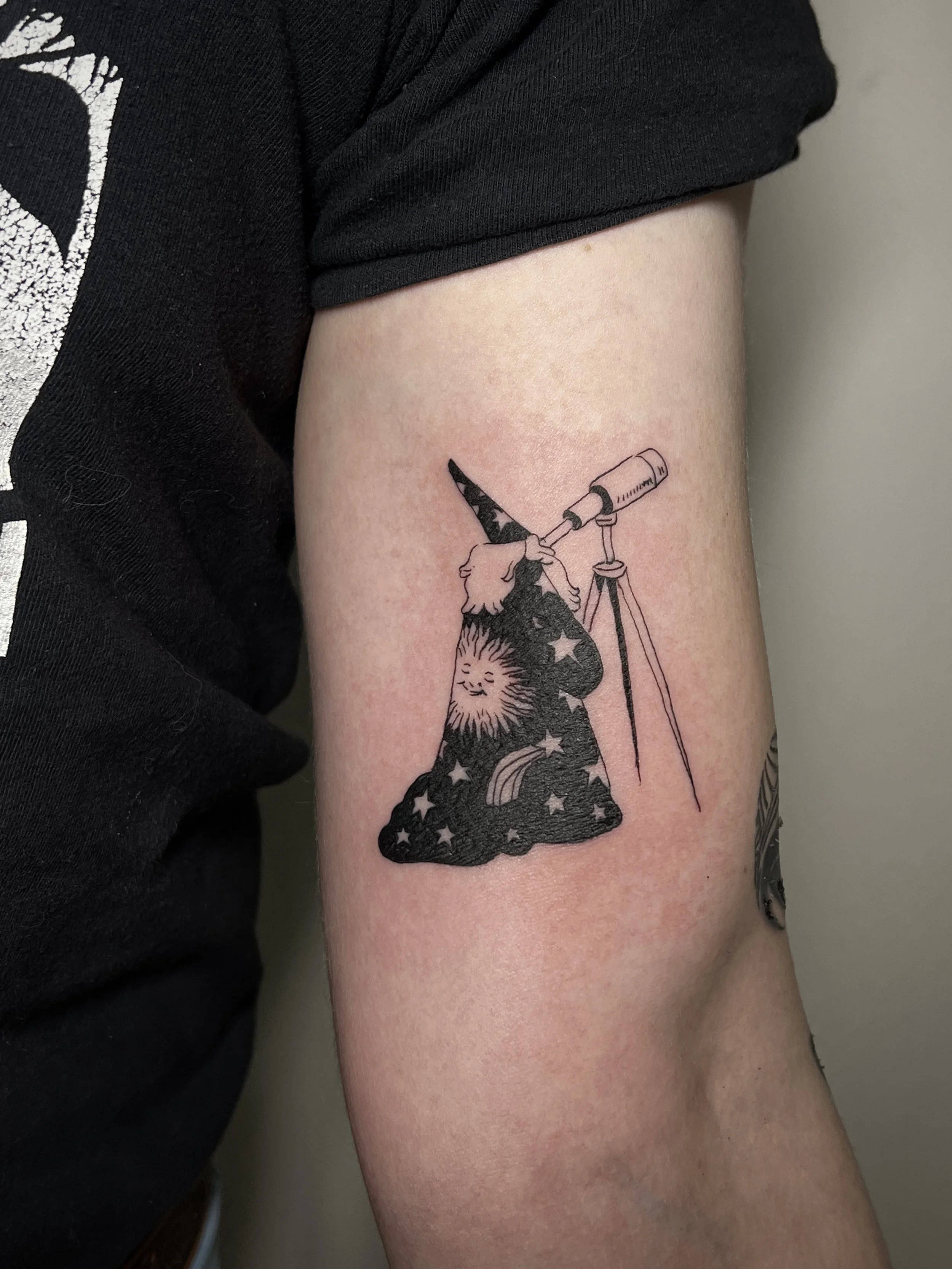 Close-up of a tattoo on a person's upper arm, featuring a wizard with a tall, pointed, starry hat peering through a telescope.