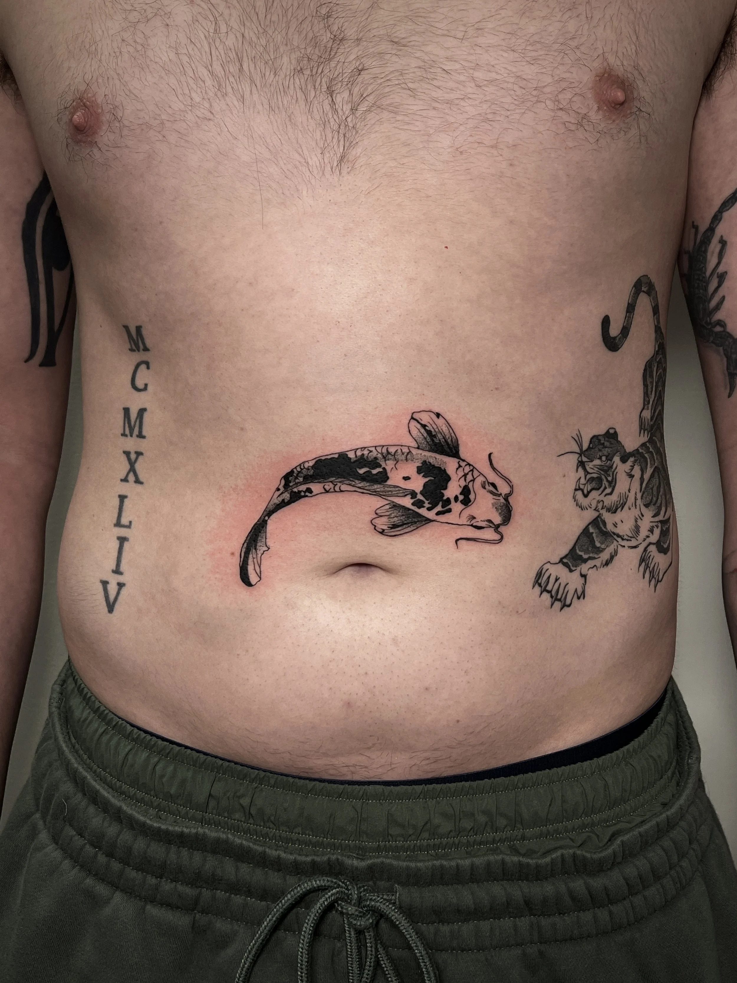 Close-up of a man's bare chest and stomach with tattoos, including a large koi fish and a tiger, and text reading 'MCMXIV' on the left side.