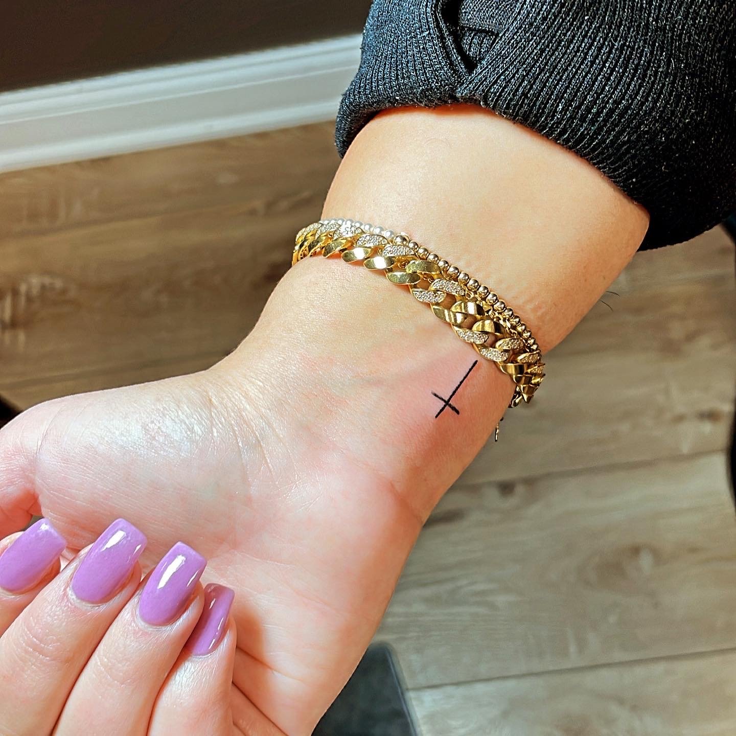 Hand with purple nails, small cross tattoo on wrist, gold bracelets on arm, wooden floor background.