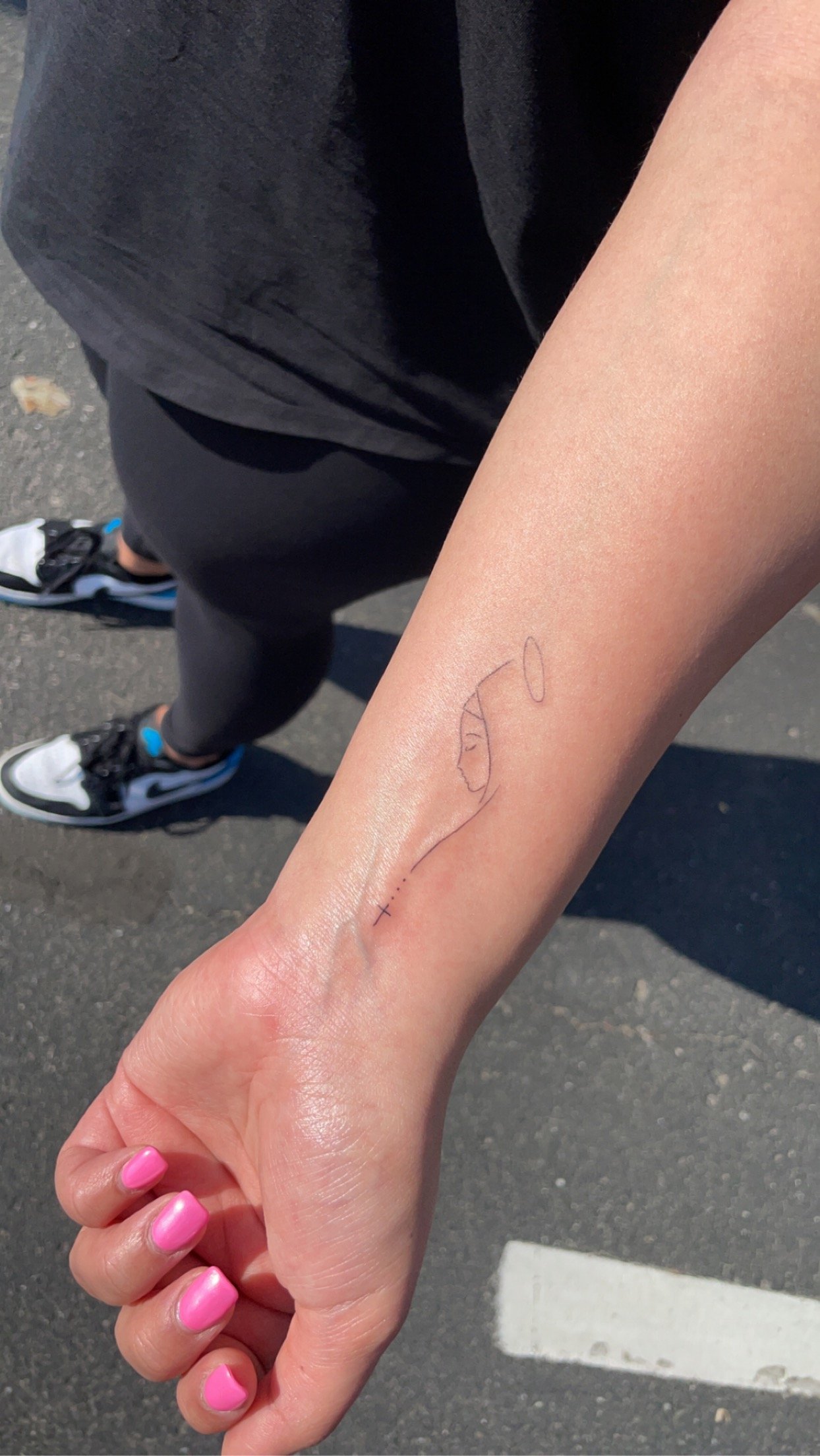 Person with a minimalistic tattoo of a woman with a halo, holding a cross on their forearm, painted pink nails, wearing black leggings and sneakers, standing on asphalt.