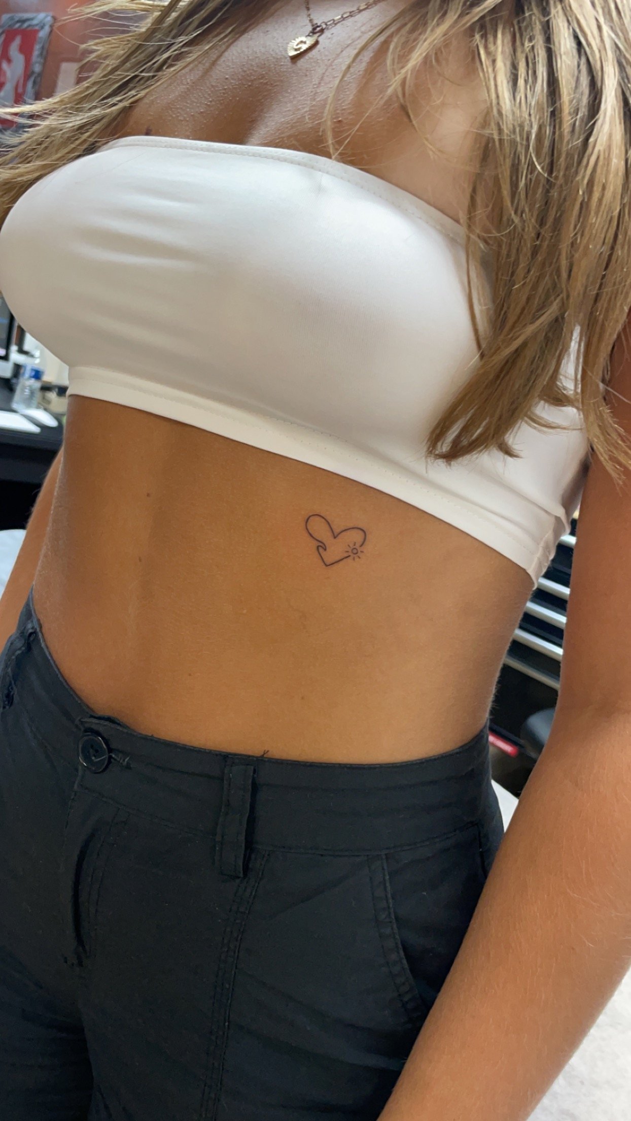 Person with a small tattoo of a heart and mouse ears near their waist, wearing a white tube top and black pants.