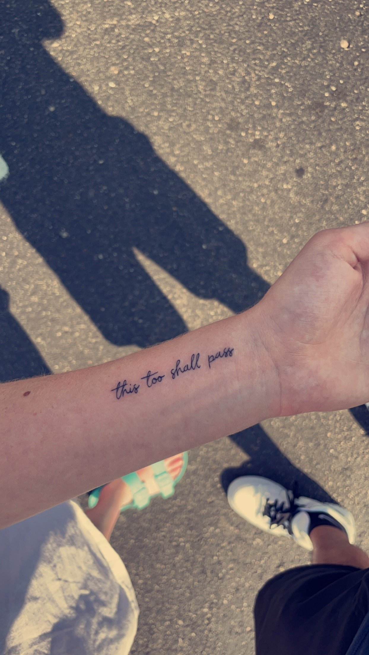 Tattoo of the phrase 'this too shall pass' on a forearm with shoes and a shadow on the pavement.