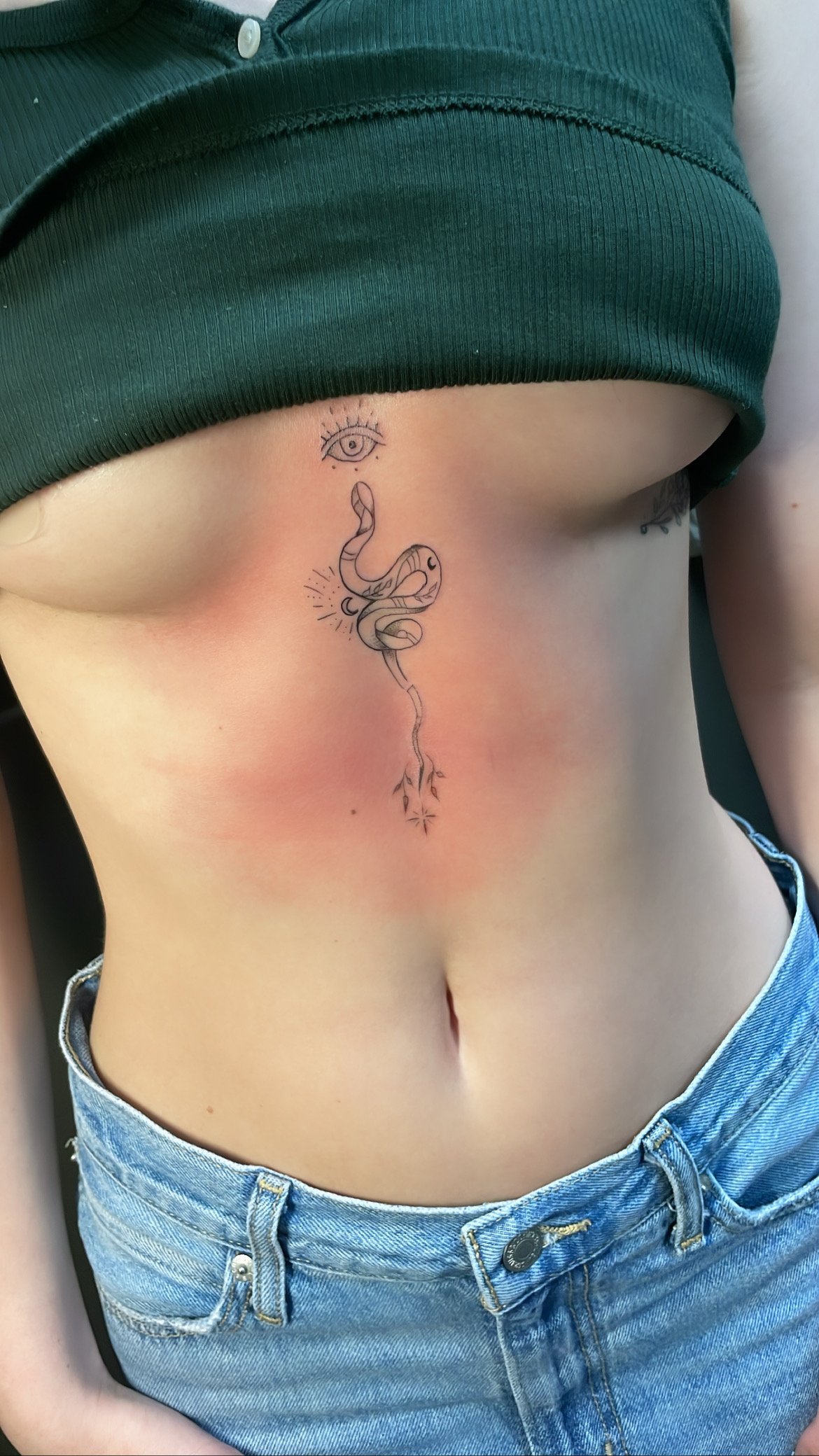 Person with a tattoo of a snake and an eye on their stomach, wearing a crop top and jeans.