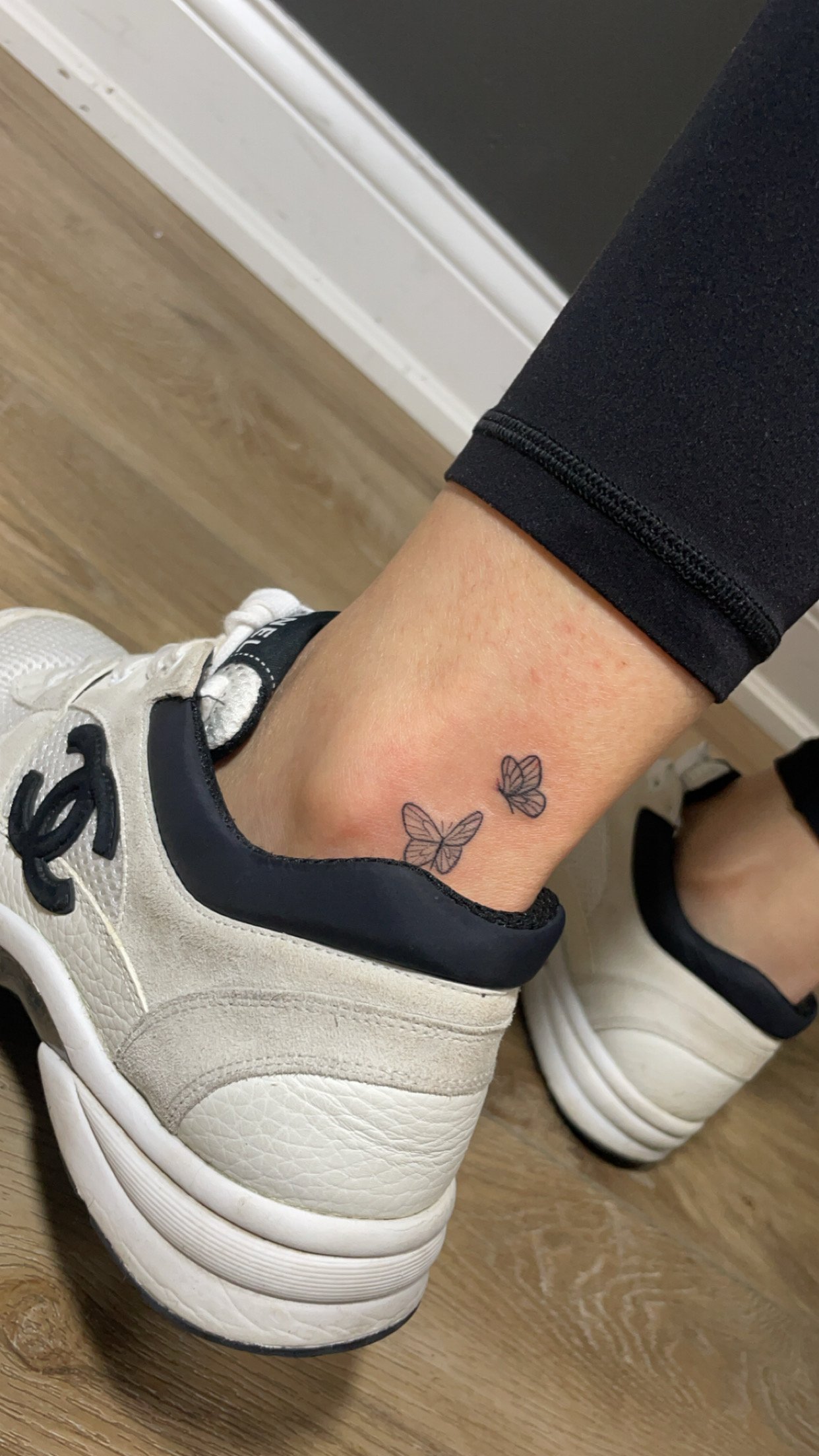 Two small butterfly tattoos on an ankle above white sneakers on a wooden floor.