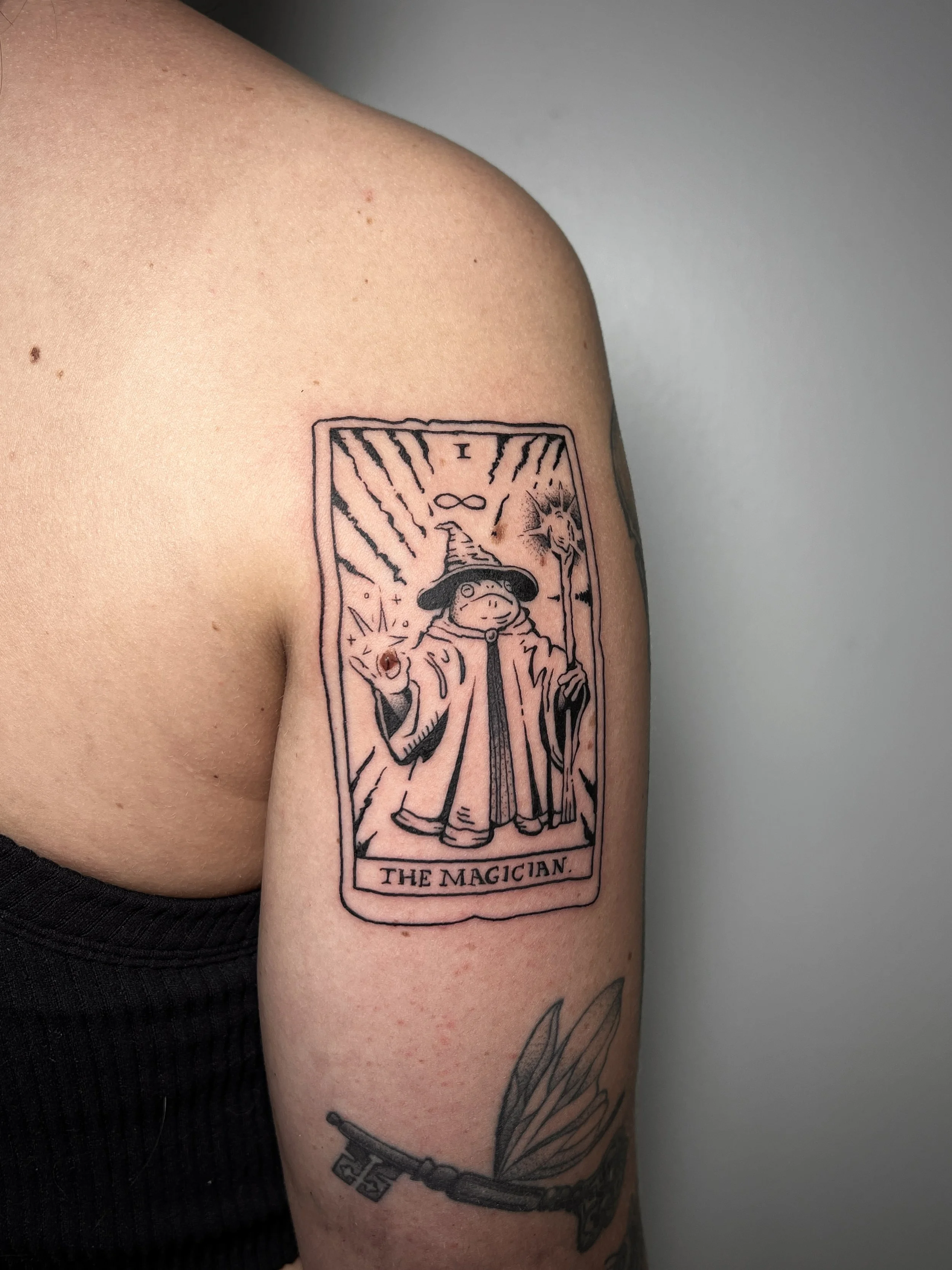 A person's upper arm with a tattoo of a tarot card labeled 'The Magician.' The card features a frog in a wizard's hat with a staff and rays behind them.