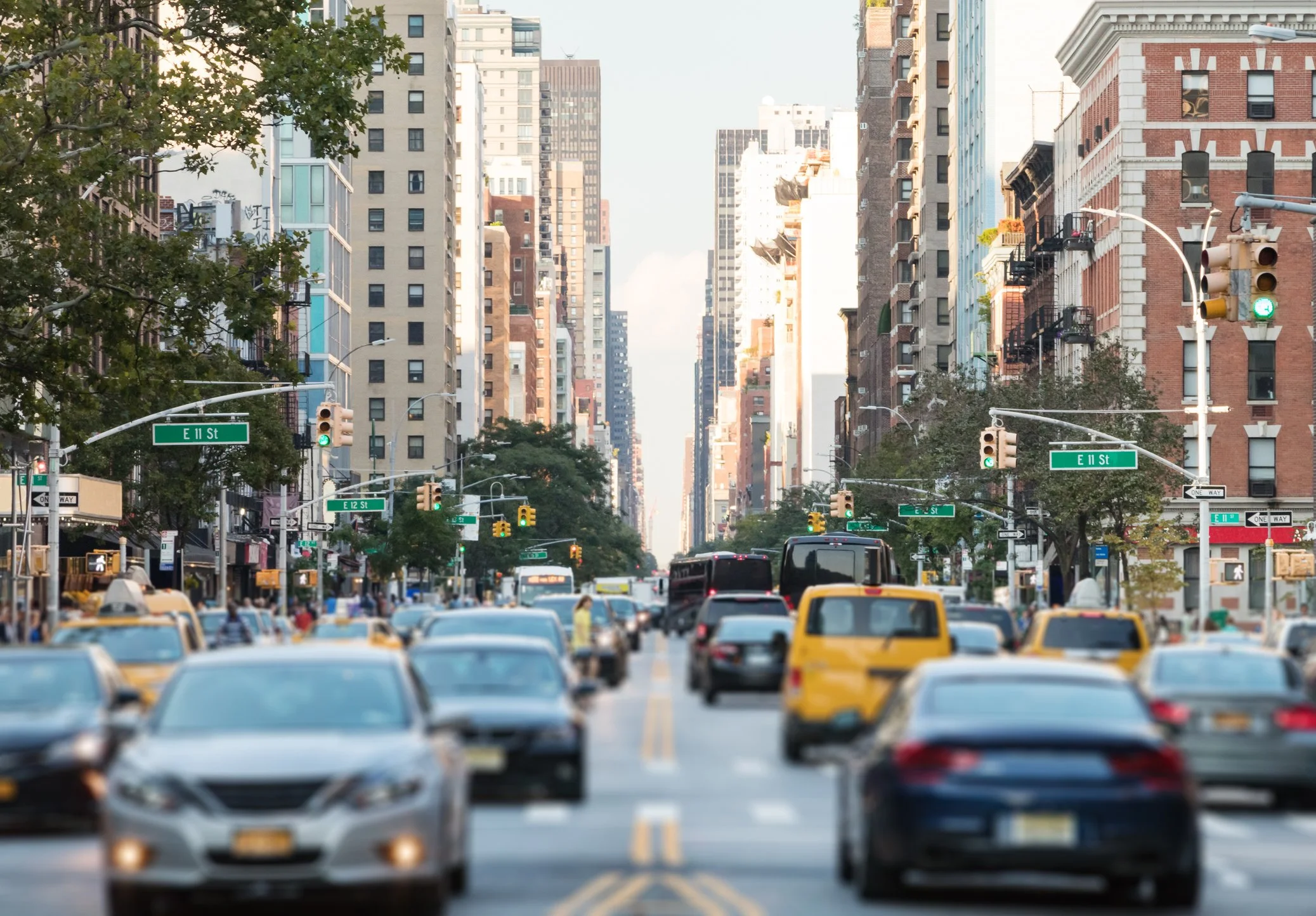 Busy city street with cars, buses, and taxis at intersection, tall buildings, and street signs indicating E 11 St and E 12 St, with trees lining the sidewalk.
