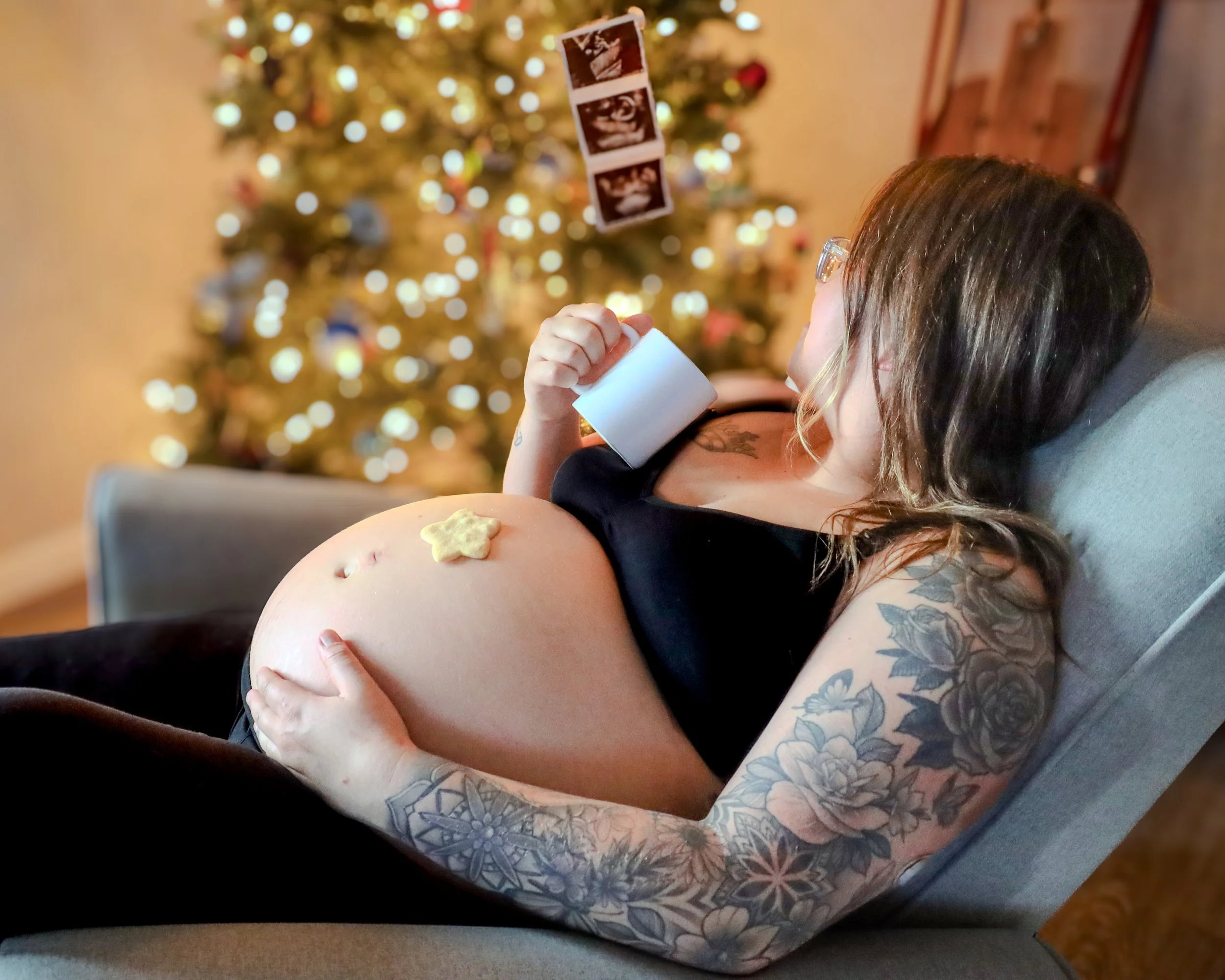Pregnant woman with tattoos holding a mug on a couch, with a Christmas tree behind her.