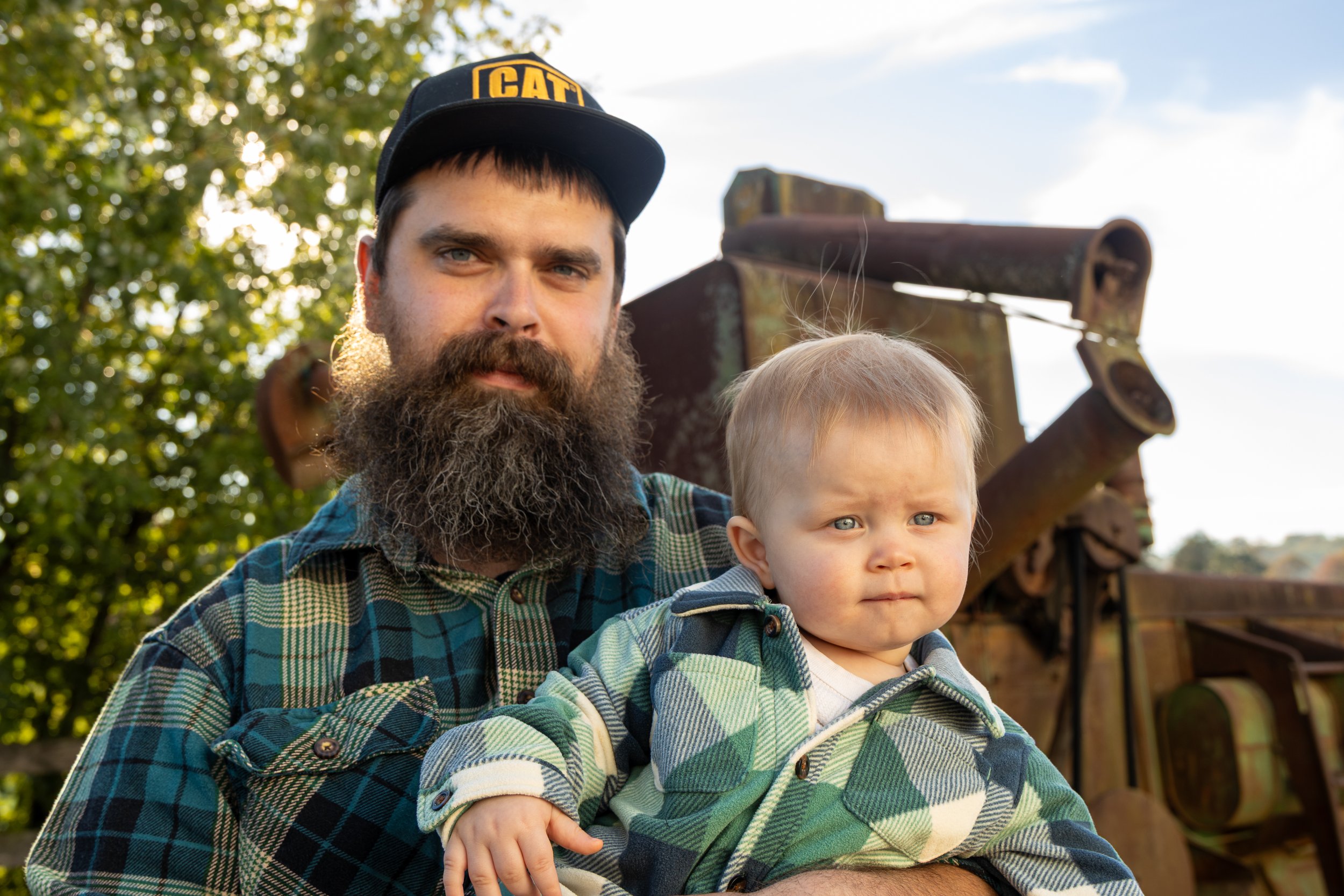 Bearded man in a cap holding a young child outdoors, with trees and sky behind them.