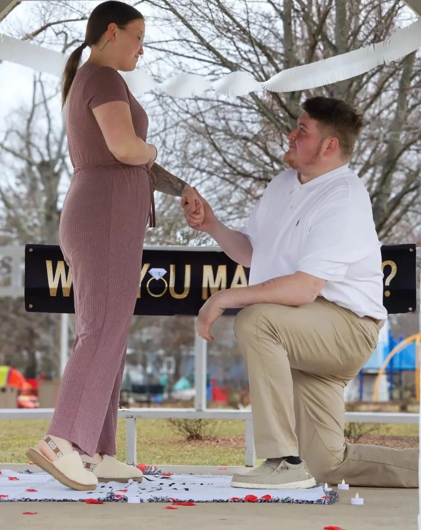 A man on one knee proposing to a woman outdoors with a 'Will You Marry Me?' sign behind them.