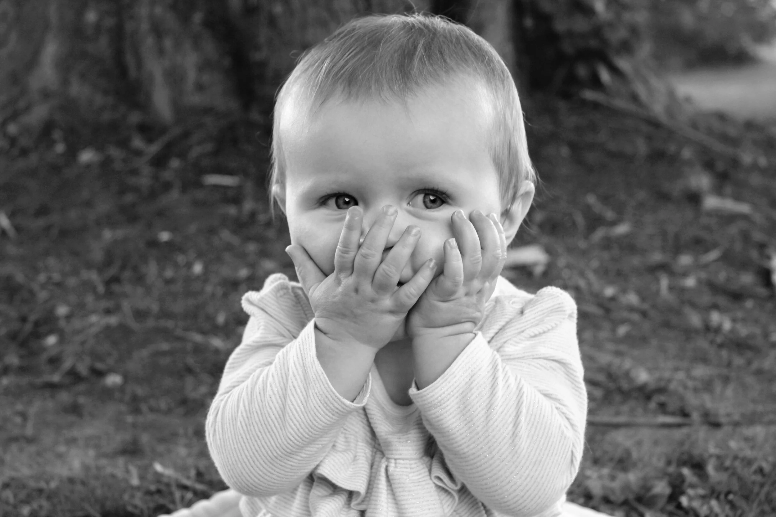 A young child with short hair covering their mouth with both hands, looking directly at the camera with wide eyes, outdoors with blurred trees in the background.