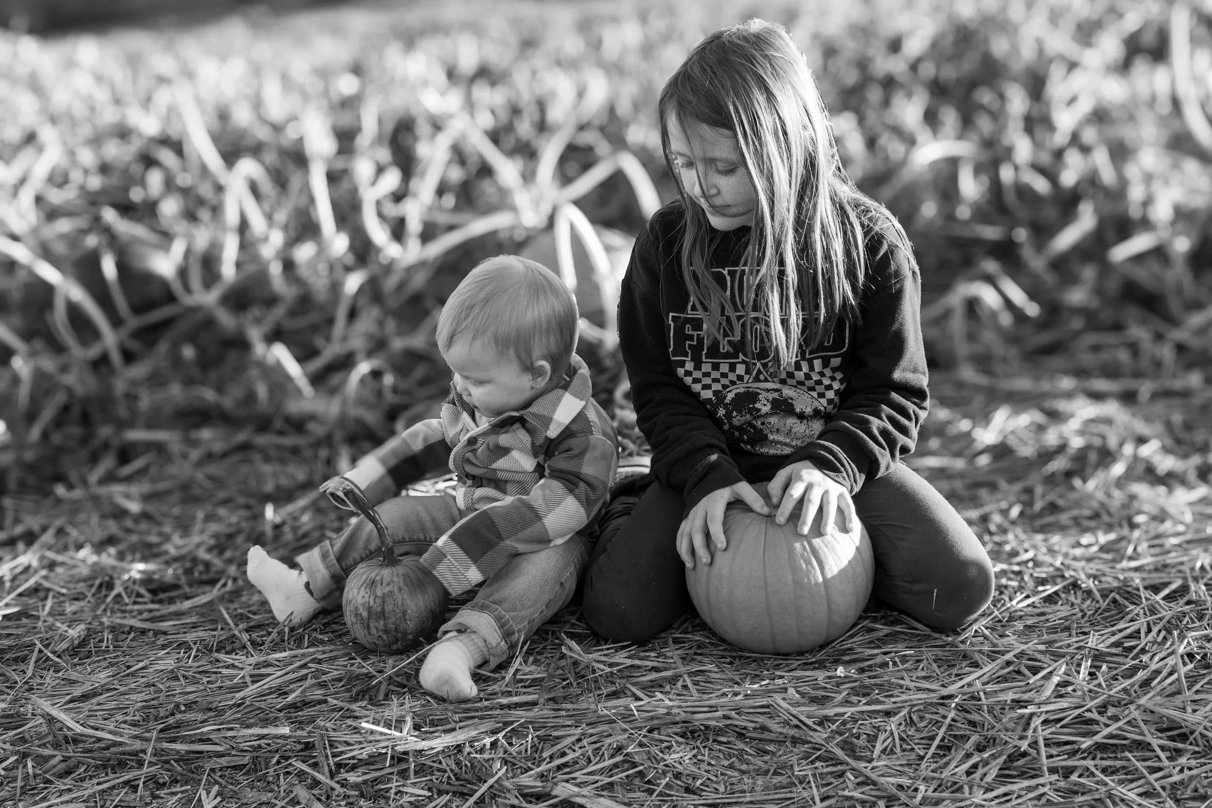 A girl and a young boy sit on the ground with pumpkins, surrounded by fallen leaves.