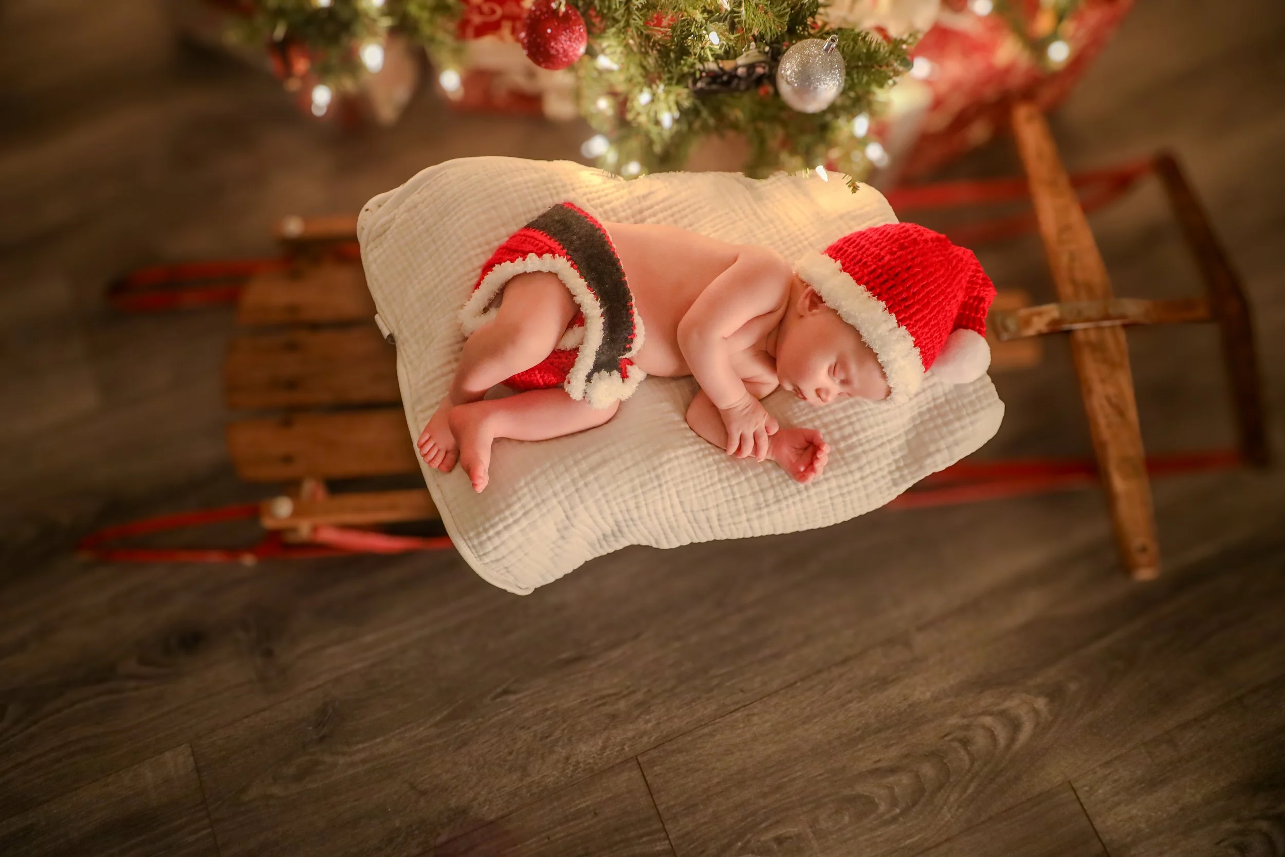 Sleeping baby on a wooden sled wearing a Santa hat, with a Christmas tree behind.