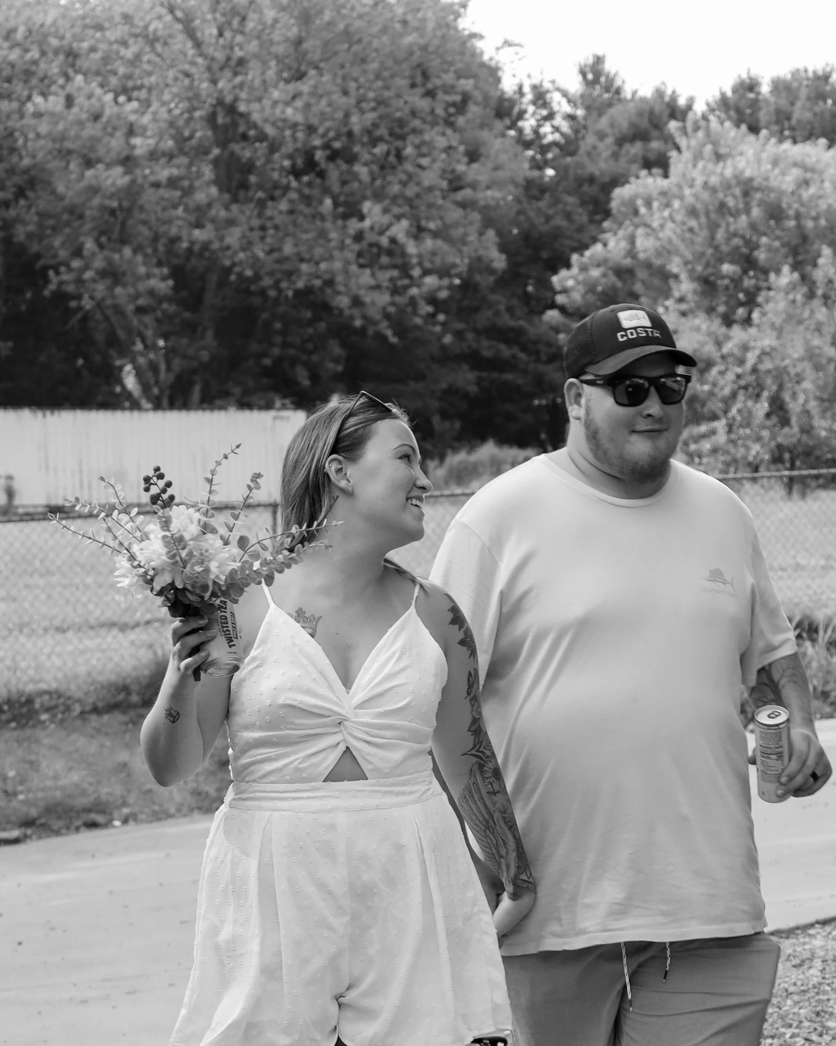 Casually dressed bride walking with bouquet, smiling at husband in sunglasses, t-shirt, and hat.