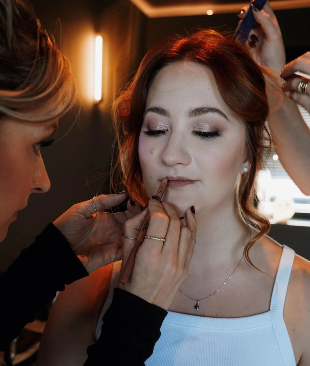 A woman getting her makeup done by a makeup artist, who is applying lip liner to her lips in a salon or makeup studio.