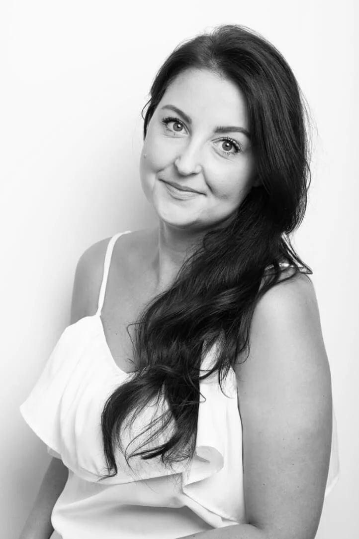 Black-and-white portrait of a woman with long, wavy dark hair, smiling softly, wearing a sleeveless white top, standing against a plain light background.