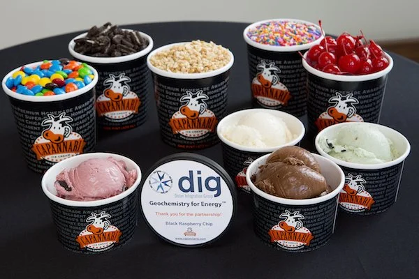 Nine cups of ice cream and toppings arranged on a black surface, with a small sign in the center that reads 'dig Geochemistry for Energy' and 'Black Raspberry Chip.' The cups have a cow logo and are branded 'Capannari.' Toppings include chocolate chips, colorful sprinkles, maraschino cherries, Oreo cookie pieces, and M&Ms.
