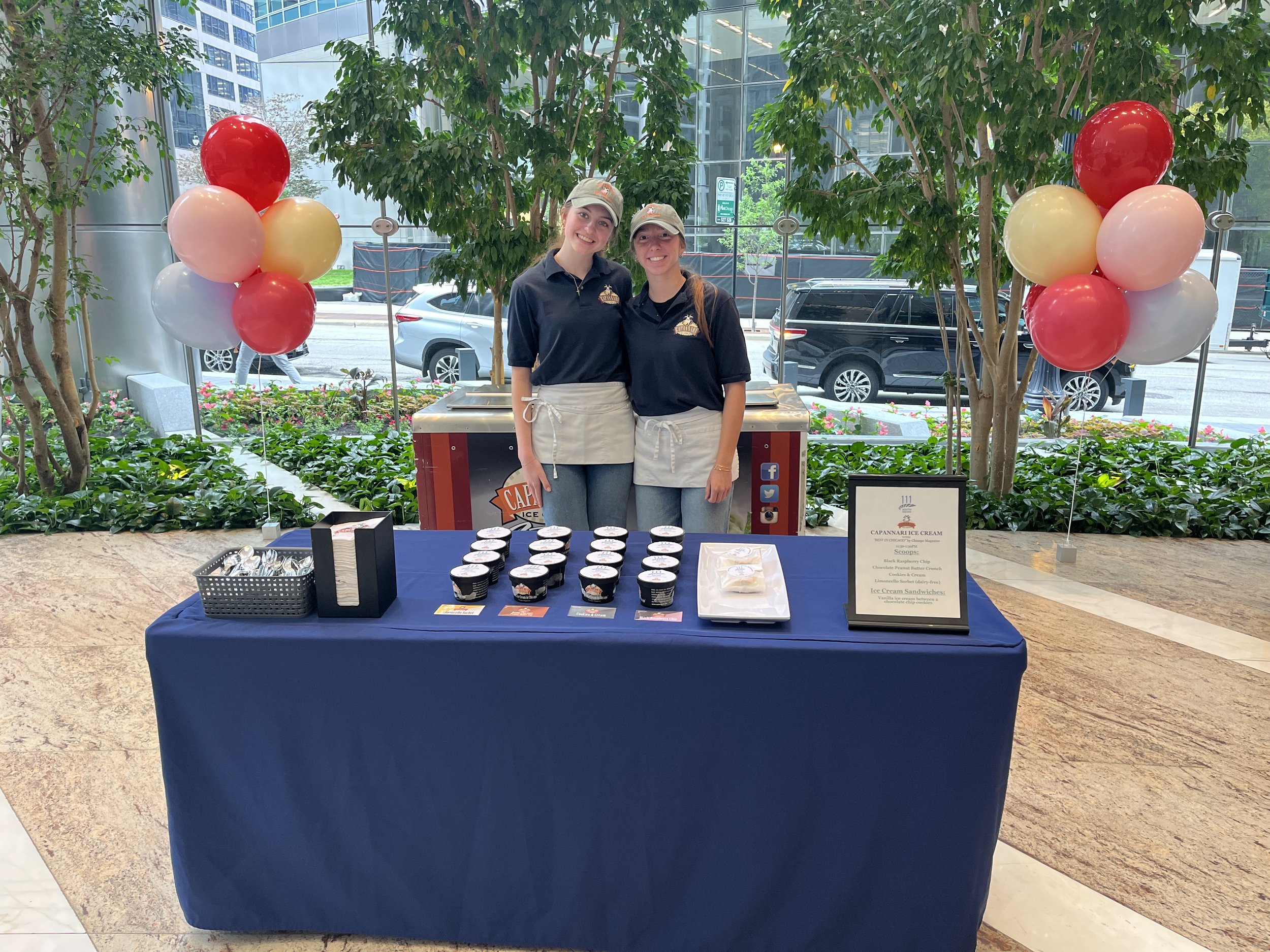 Two women standing behind a table at an ice cream stand, smiling at the camera, with balloons on either side. The table is covered with a dark blue cloth and displays small cups of ice cream and a sign. The background includes plants, trees, and a vi
