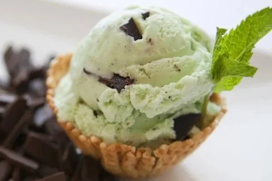 A scoop of green mint chocolate chip ice cream in a waffle cup, garnished with a mint leaf, with chocolate shavings around it.